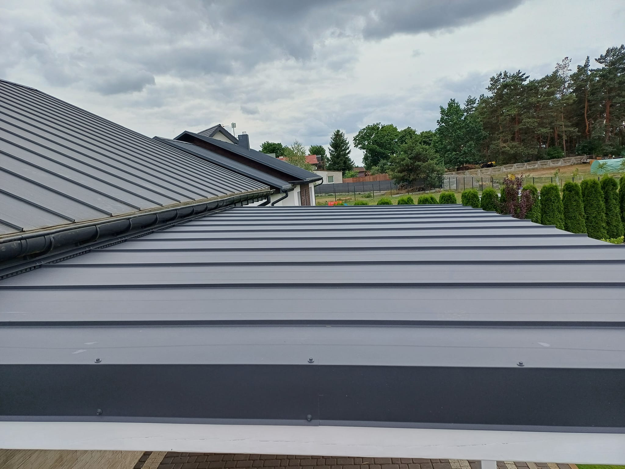 Nowoczesne, szare pokrycie dachowe z blachy na domu jednorodzinnym, widoczne z góry, z rynnami i elementami mocującymi, z widokiem na ogród i drzewa w tle pod pochmurnym niebem.