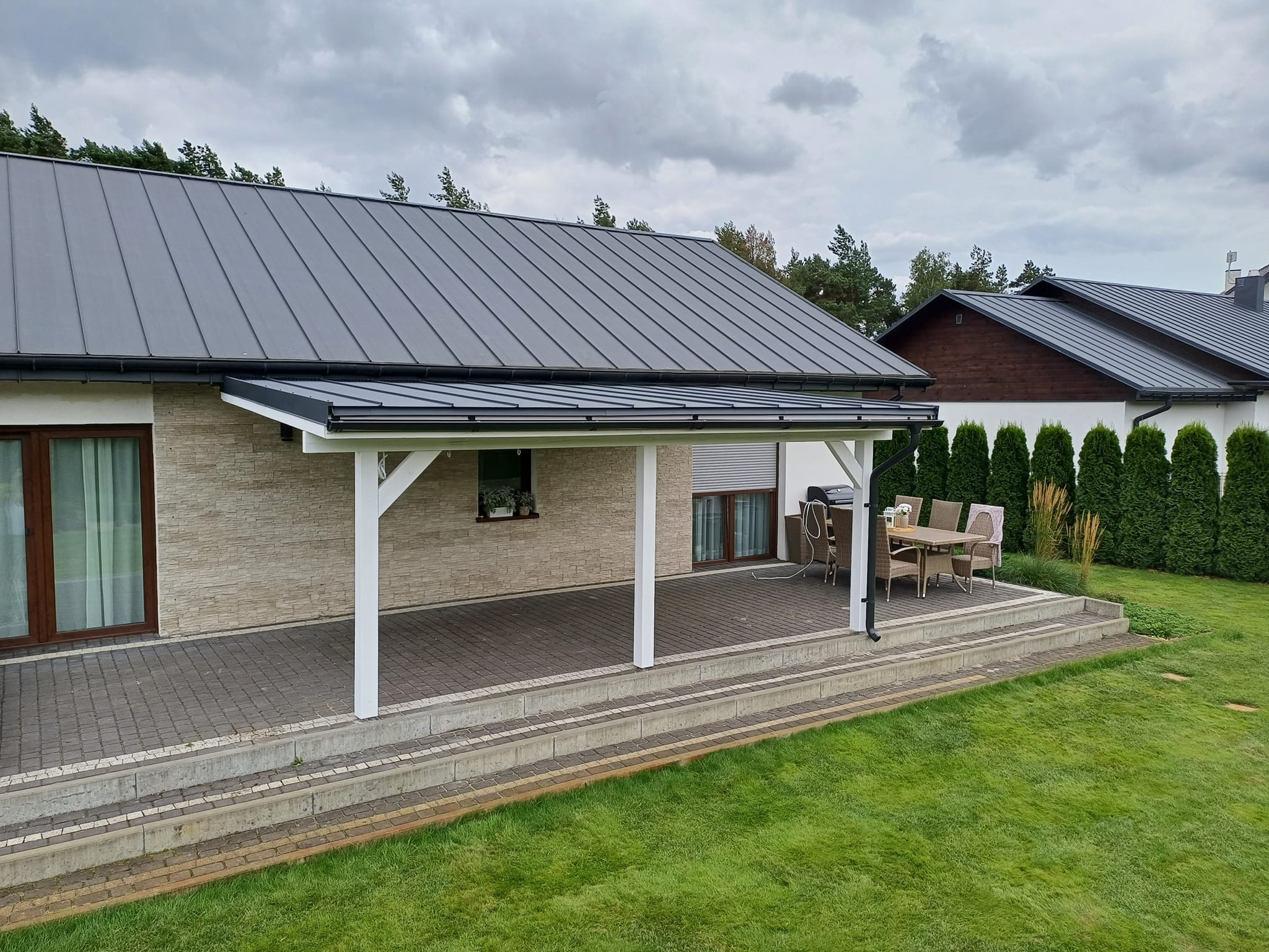 Zadaszony taras z białymi słupami, wyłożony kostką brukową, z widocznym zestawem mebli ogrodowych i fragmentem zadbanego trawnika.