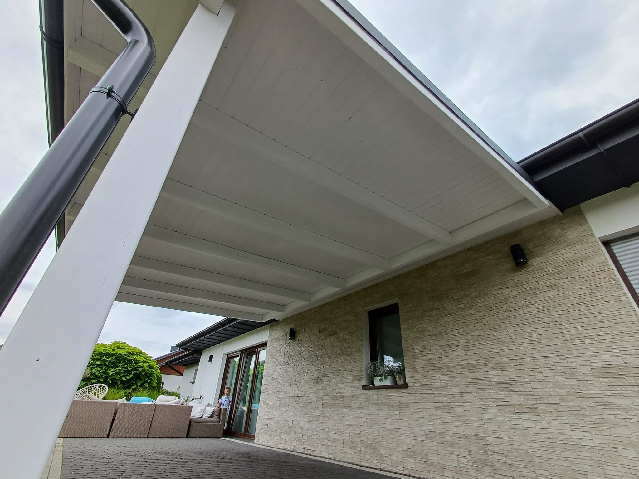 Zadaszona przestrzeń tarasowa z białym sufitem, widok z dołu, z widocznym fragmentem elewacji z kamienia dekoracyjnego i meblami ogrodowymi w tle.