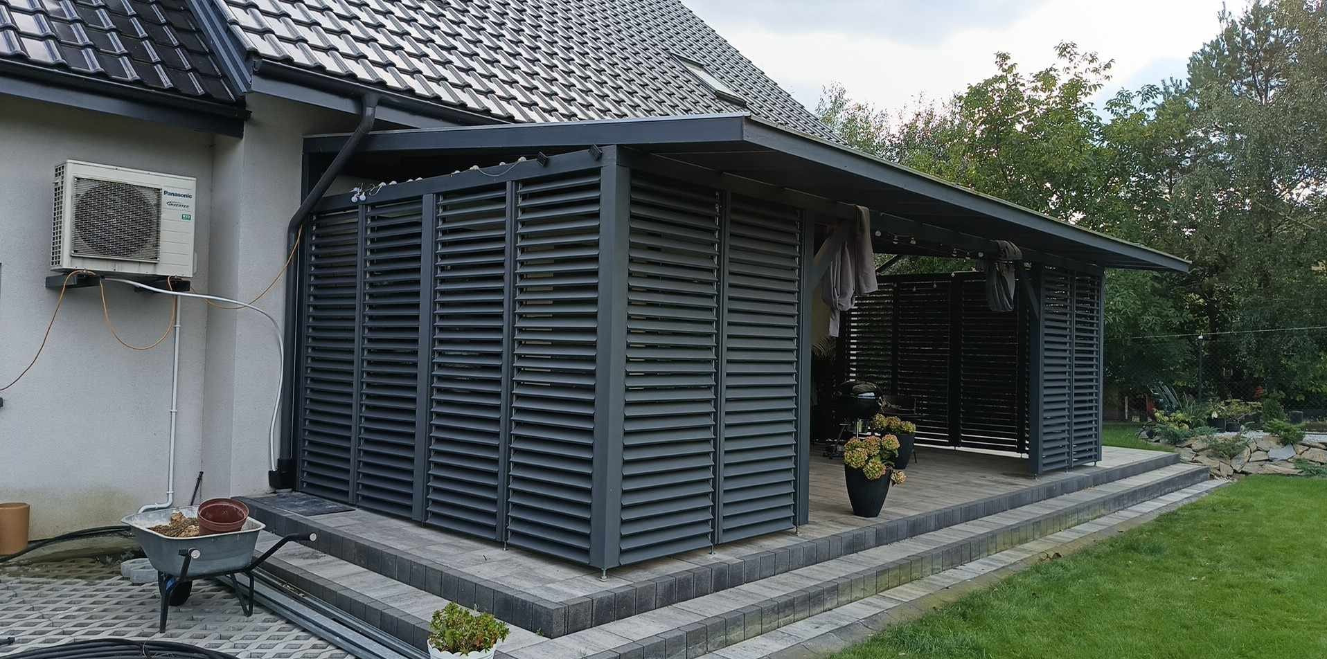 Nowoczesna pergola z szarymi żaluzjami, przylegająca do domu z klimatyzatorem, widok z boku na taras z donicami, w tle zieleń ogrodu.