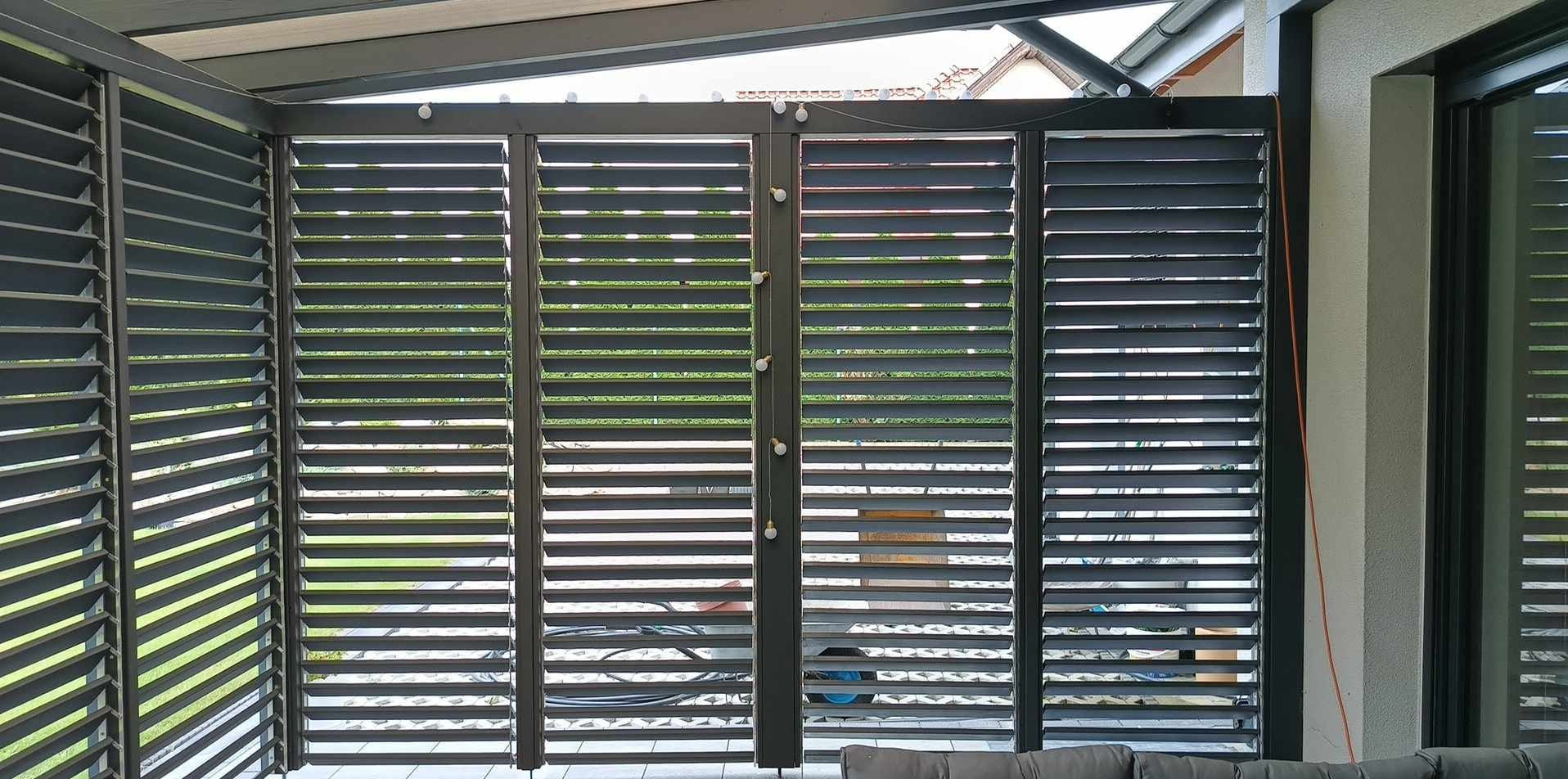 Nowoczesna pergola z regulowanymi lamelami w kolorze antracytowym, oświetlona dekoracyjnymi lampkami, z widokiem na zieleń ogrodu w tle.