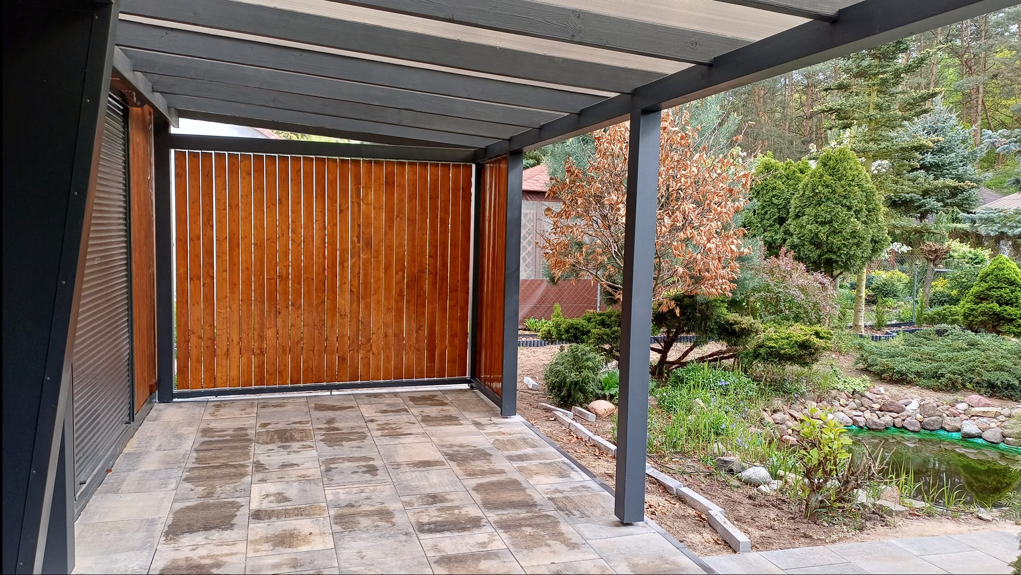 Zadaszona przestrzeń z drewnianą ścianą z pionowych desek i szarą konstrukcją, wyłożona płytkami, z widokiem na ogród z oczkiem wodnym.