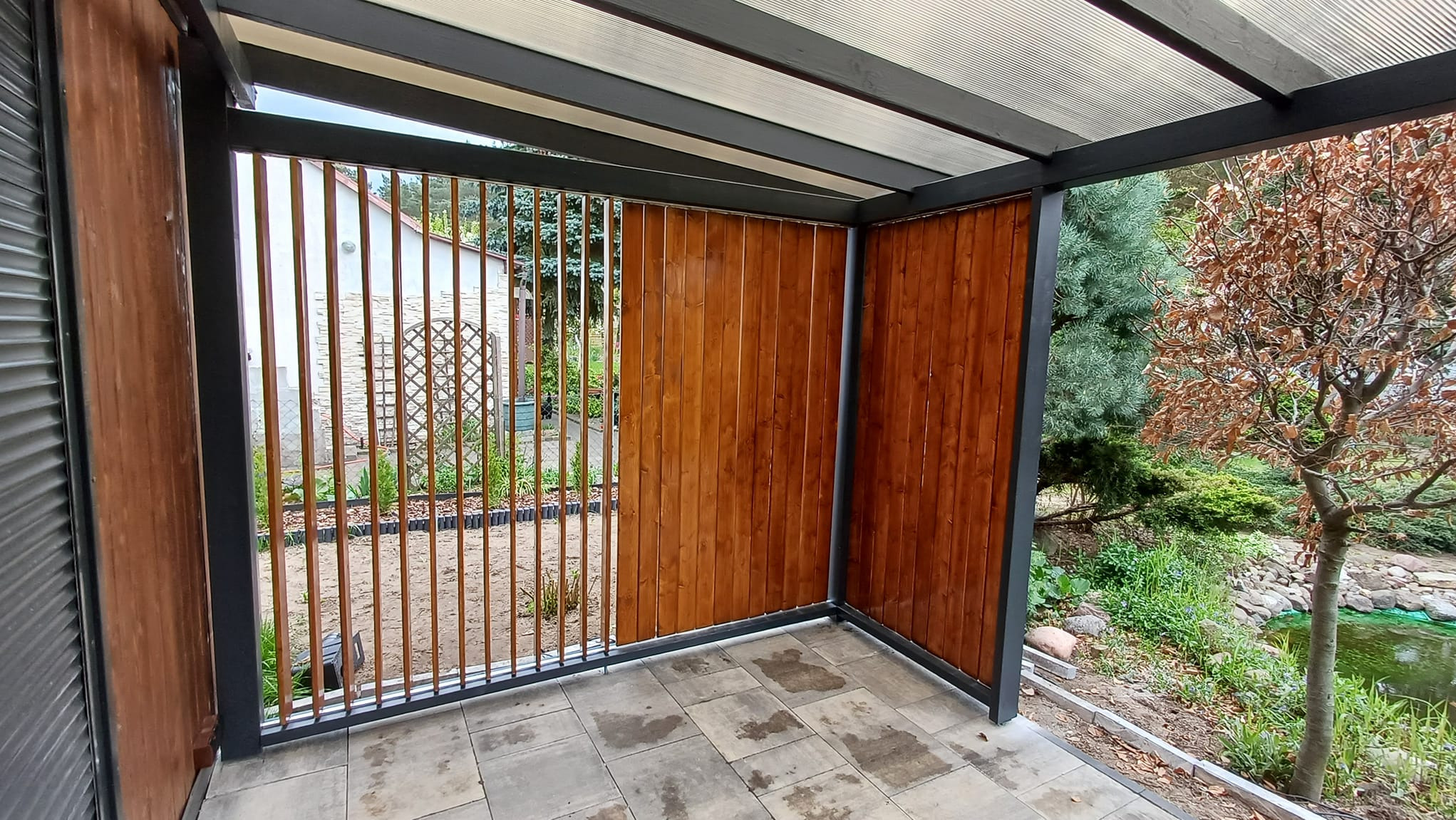 Zadaszona konstrukcja z drewnianymi panelami, tworząca osłonę od wiatru i słońca, z widokiem na ogród z oczkiem wodnym i drzewem o brązowych liściach.