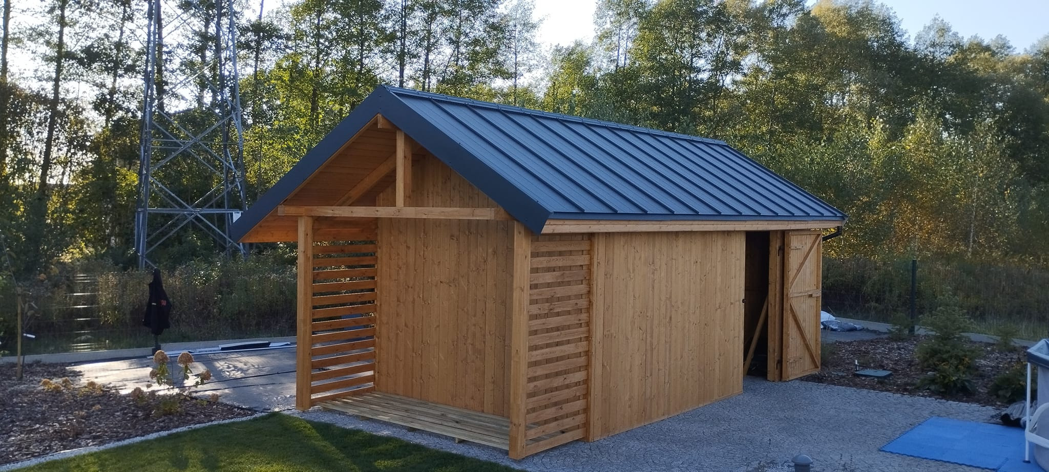 Drewniana altana z szarą, metalową dachówką, częściowo osłonięta ażurowymi panelami, z otwartymi drzwiami, na tle zieleni i słupa wysokiego napięcia.
