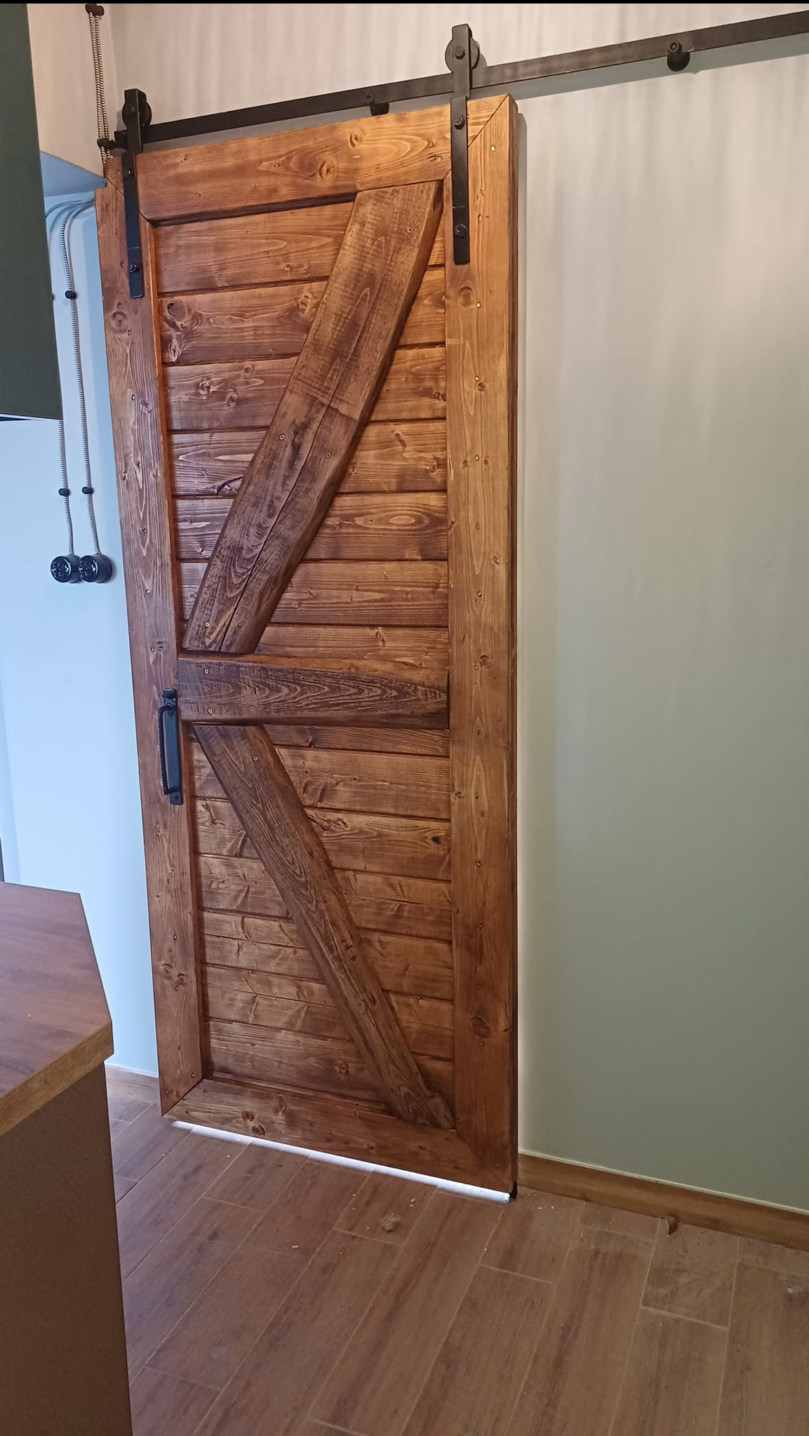 Rustykalne, przesuwne drzwi drewniane w stylu stodoły z czarnymi okuciami na szynie, widoczne w jasnym wnętrzu z drewnopodobną podłogą.