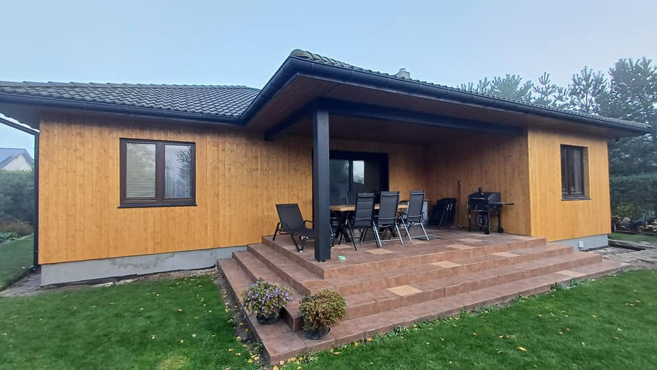 Zadaszony taras z brązowych płytek, drewniana elewacja domu, czarne słupy podtrzymujące zadaszenie, stół z krzesłami, grill na zewnątrz.