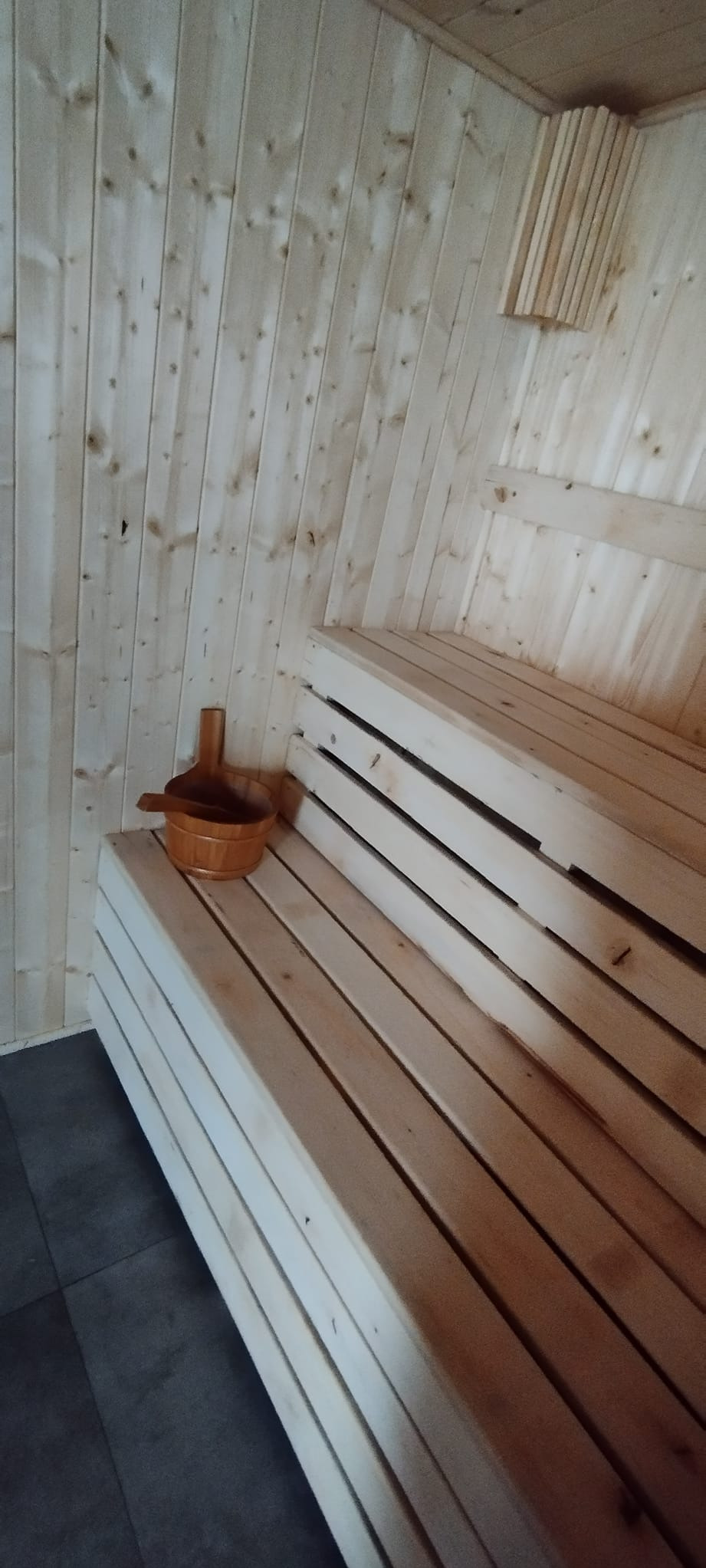 Wnętrze sauny wykończone jasnym drewnem, z drewnianymi ławami i cebrzykiem z chochlą na jednej z nich.