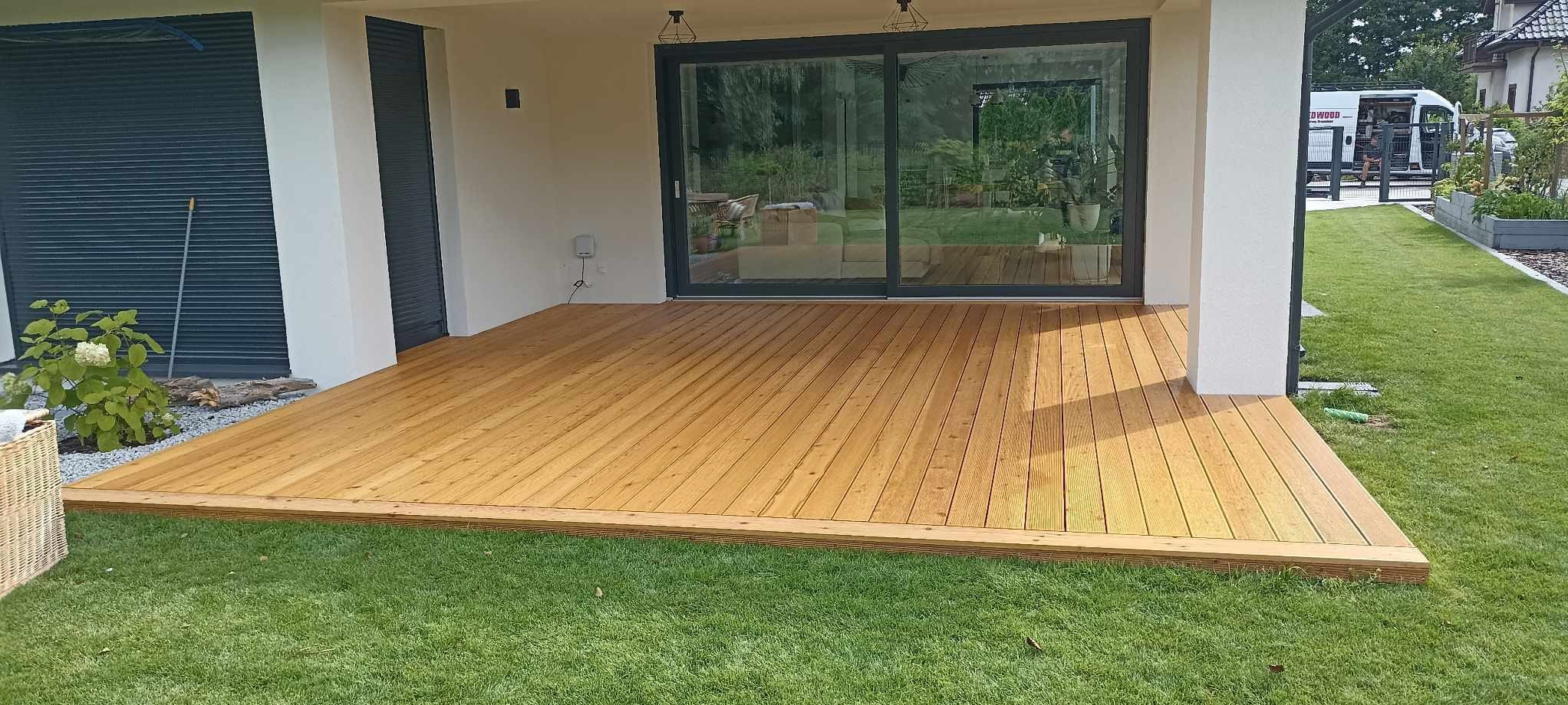 Nowoczesny taras z desek w kolorze jasnego drewna, przylegający do domu z dużymi przesuwnymi drzwiami, otoczony zadbanym trawnikiem.