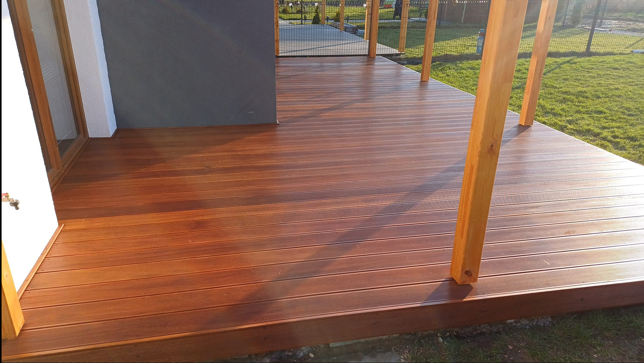 Nowoczesny taras z desek kompozytowych w ciepłym odcieniu brązu, z widocznymi drewnianymi słupami konstrukcyjnymi i fragmentem trawnika w tle.