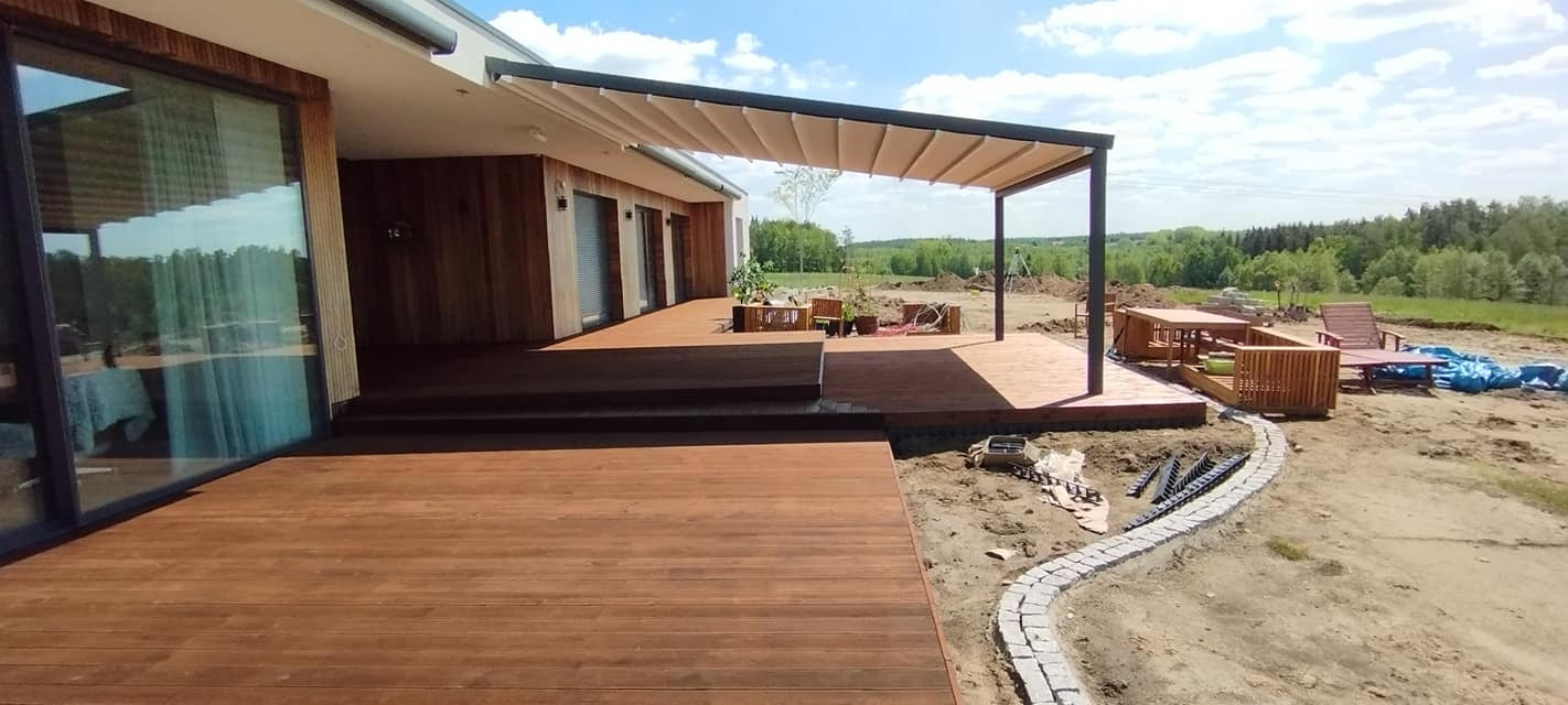Nowoczesny taras drewniany z zadaszeniem pergolą w kolorze antracytowym, połączony z drewnianą elewacją domu, widoczne prace wykończeniowe w ogrodzie.