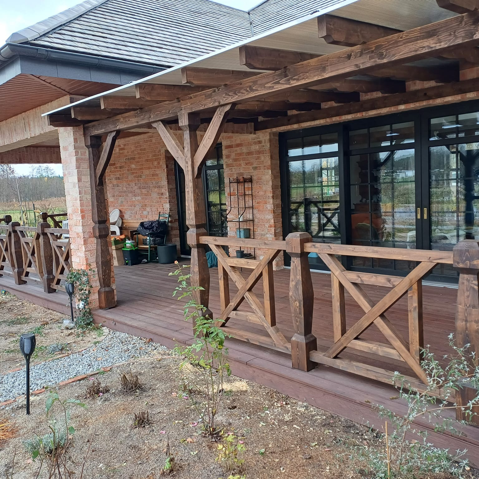 Zadaszony taras z drewnianą balustradą w stylu rustykalnym, widok z ogrodu, ceglana elewacja domu.