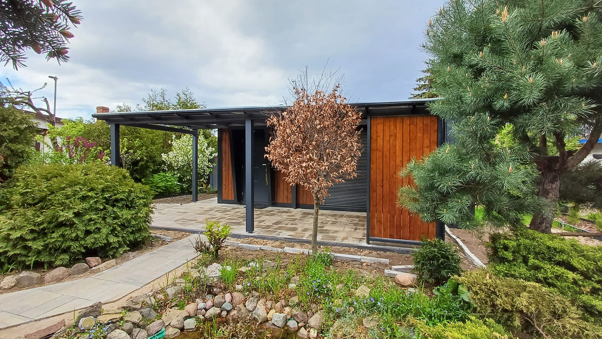 Nowoczesna altana z drewnianymi panelami i zadaszeniem, otoczona zielenią ogrodu z oczkiem wodnym i kamienną ścieżką.