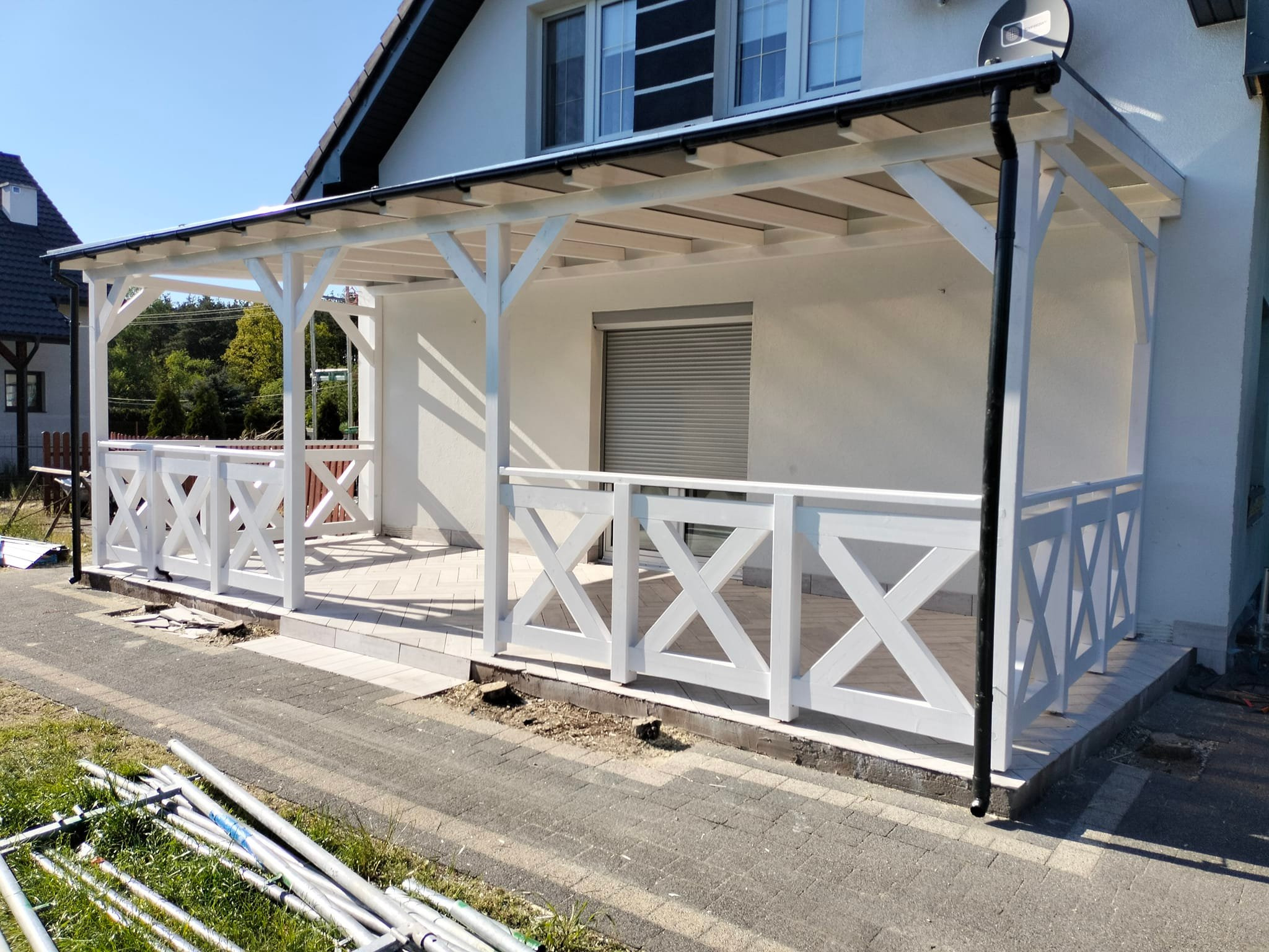 Biała drewniana pergola z balustradą krzyżową przy białym domu, widoczne rury montażowe na trawie i fragment chodnika.