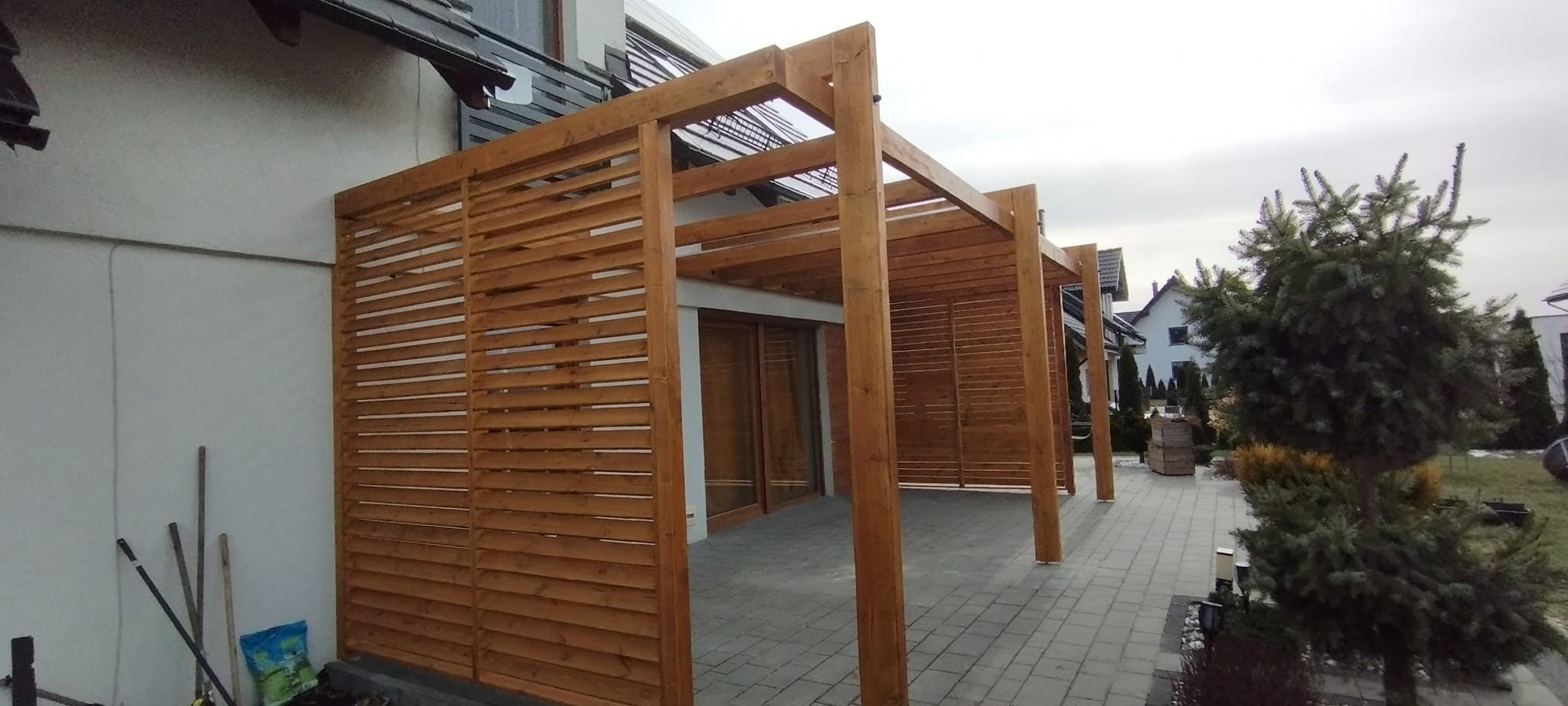 Drewniana konstrukcja z lamelami, przylegająca do budynku, tworząca zadaszone przejście. Widoczne elementy konstrukcyjne z naturalnego drewna oraz szare płytki chodnikowe. W tle domy jednorodzinne...