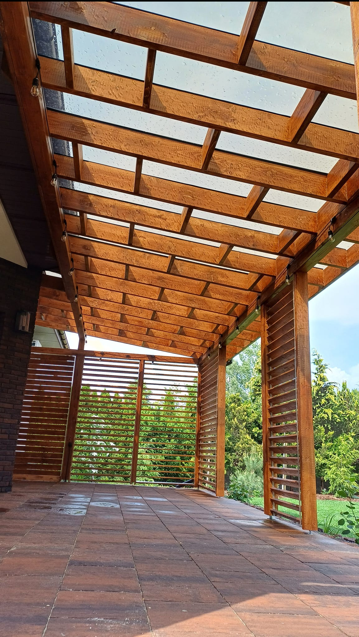 Zadaszona konstrukcja z drewna w Zławieś Wielka, z przezroczystym dachem i drewnianymi lamelami po bokach, oświetlona wiszącymi żarówkami, widok z dołu na brązową kostkę brukową.