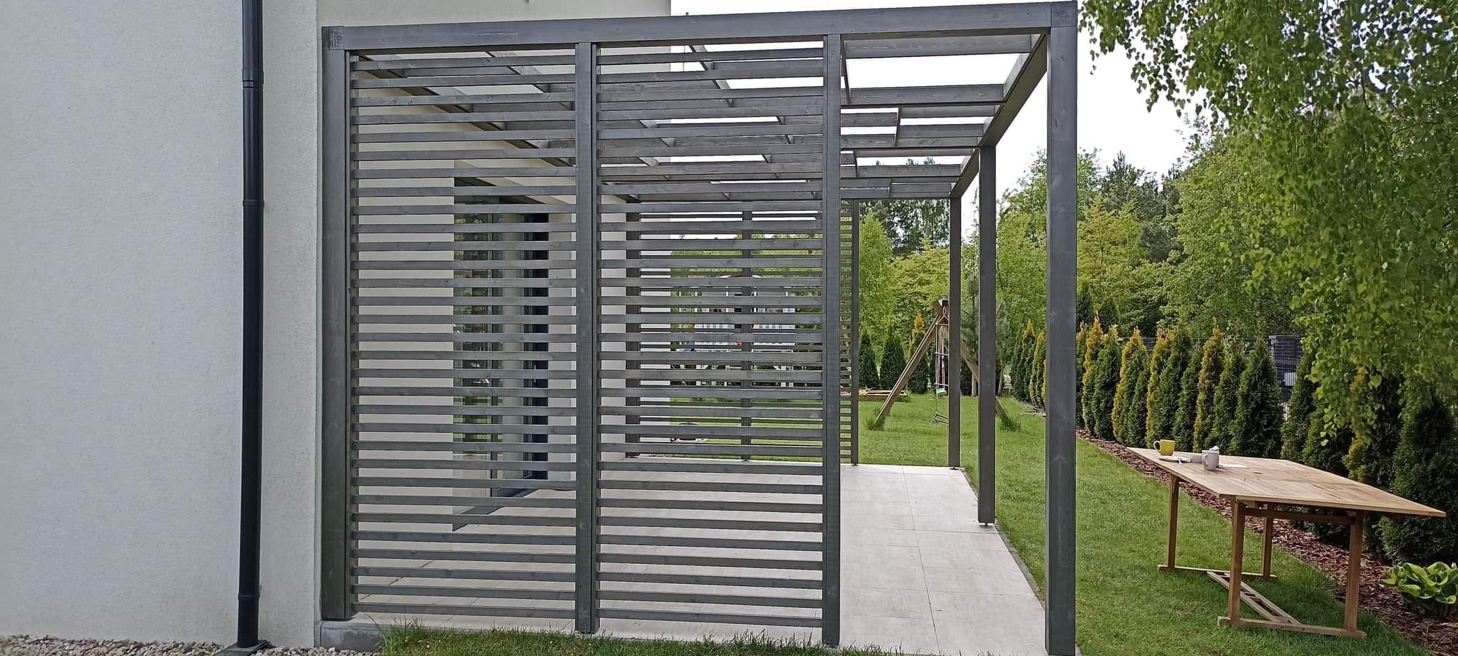 Nowoczesna, szara konstrukcja z drewna z poziomymi szczeblami, tworząca częściowo zadaszone patio z widokiem na ogród z drzewami i stołem.
