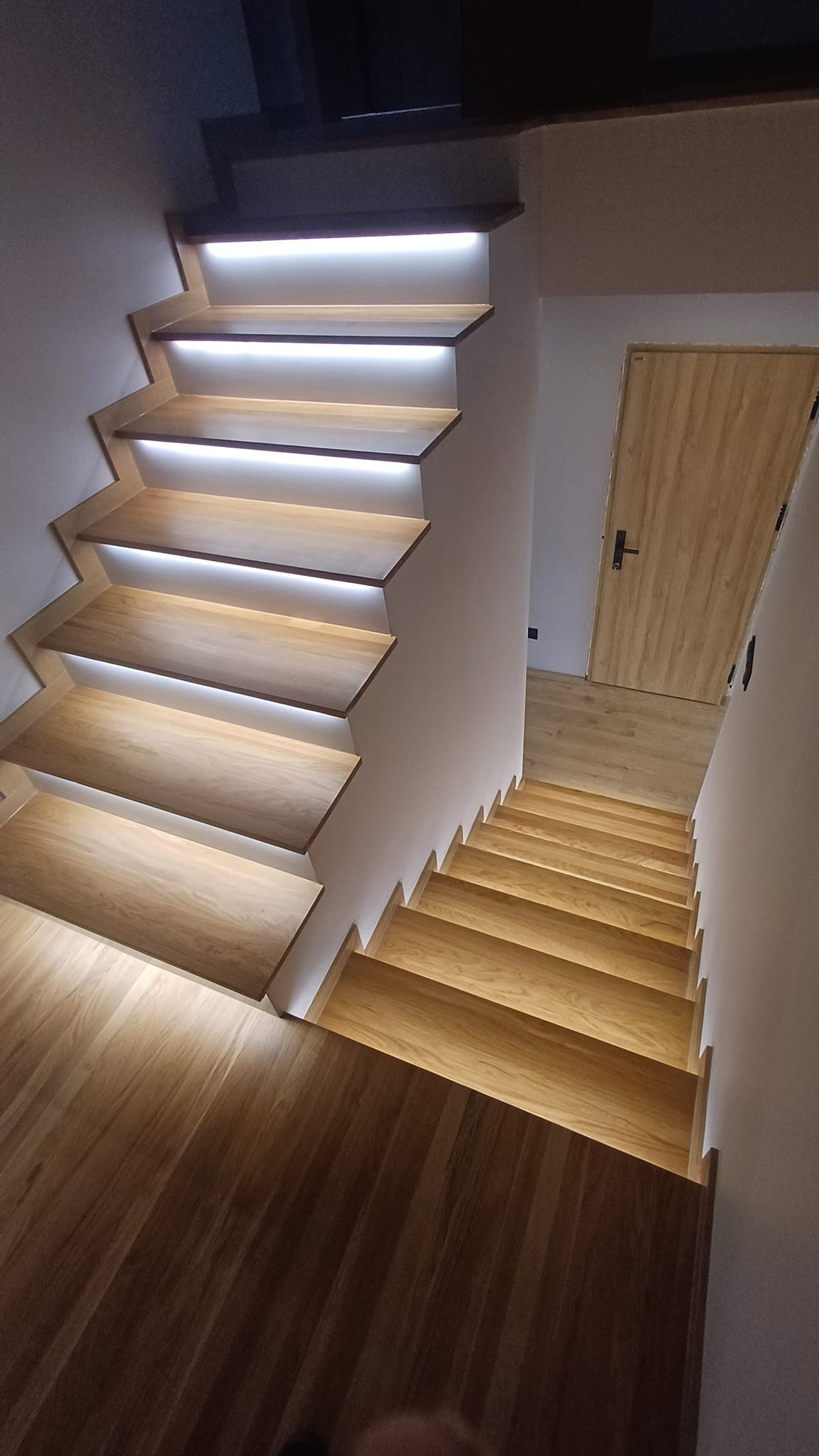 Nowoczesne schody drewniane z podświetleniem LED, widok z góry na dół, jasne drewno, białe ściany, drzwi w tle.