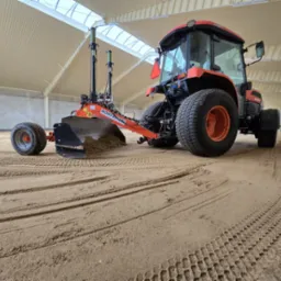 Pomarańczowy traktor Kubota z laserowym systemem niwelacyjnym, wyrównujący piasek w hali o białym, falistym suficie i ścianach. Widoczne ślady opon na piasku.