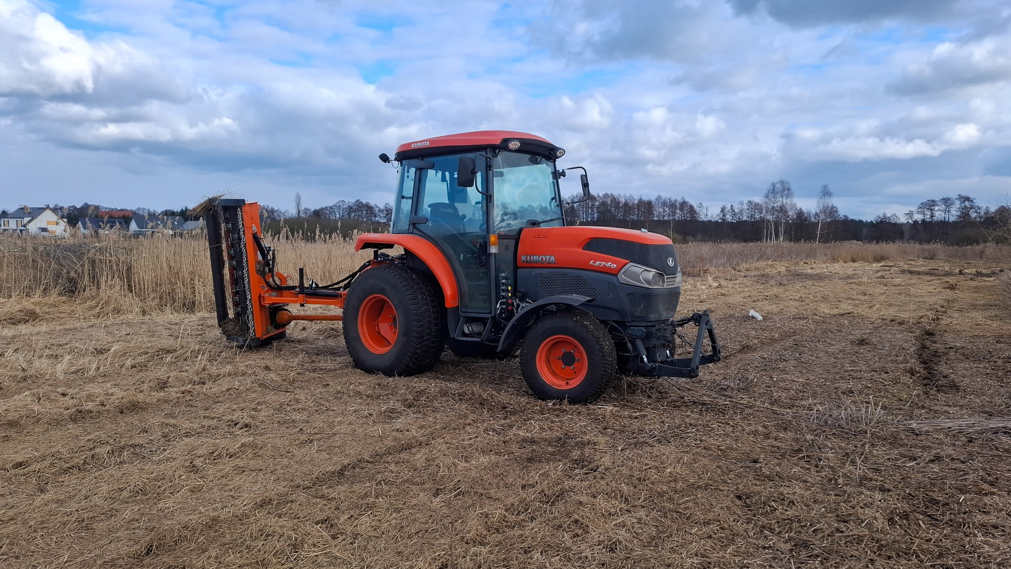 Pomarańczowy traktor Kubota L5740 z zamontowaną kosiarką listwową na polu z wysoką, suchą trawą; w tle widoczne zabudowania mieszkalne i drzewa.