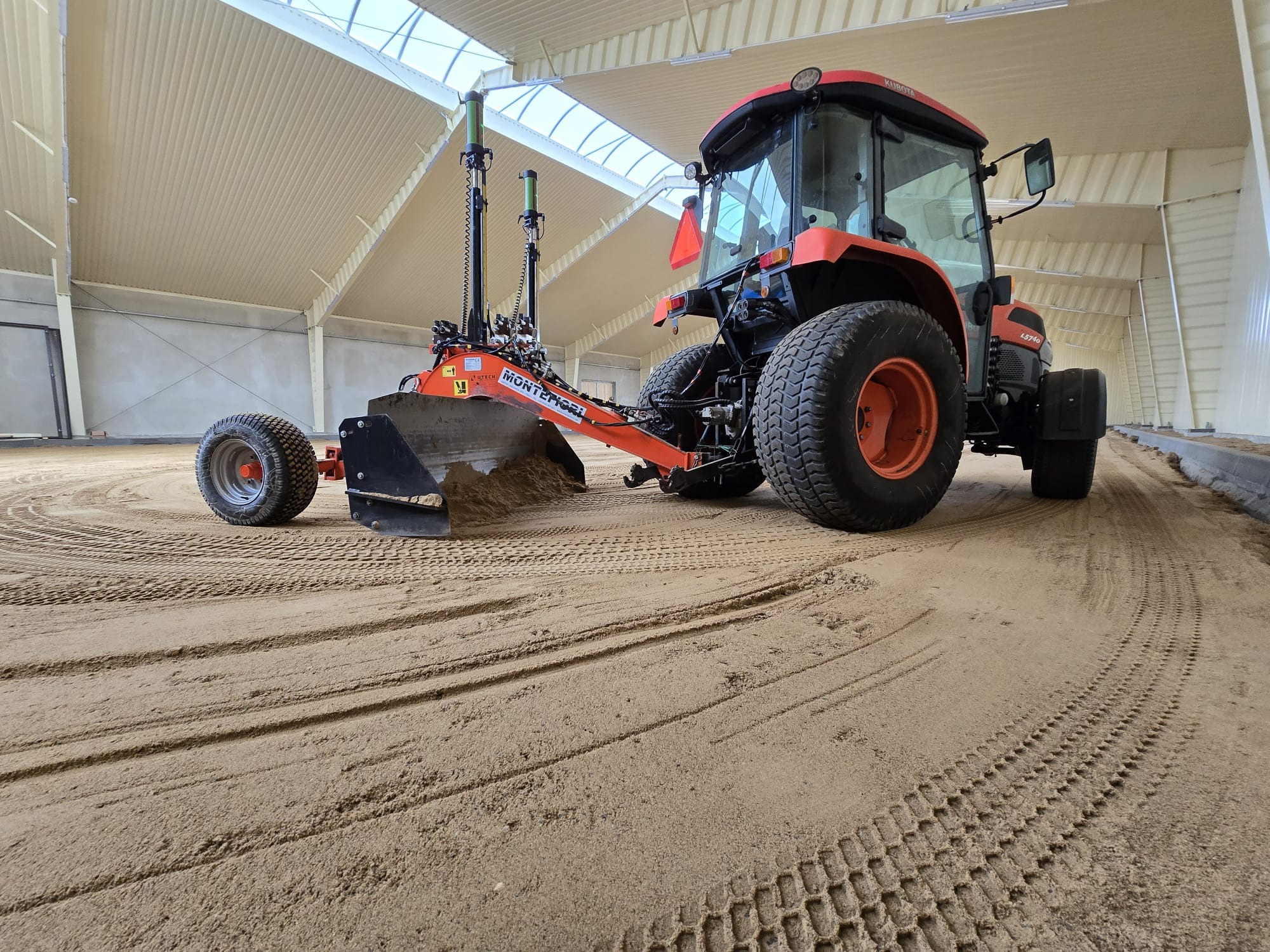 Pomarańczowy traktor Kubota z laserowym systemem niwelacyjnym, wyrównujący piasek w hali o białym, falistym suficie i ścianach. Widoczne ślady opon na piasku.