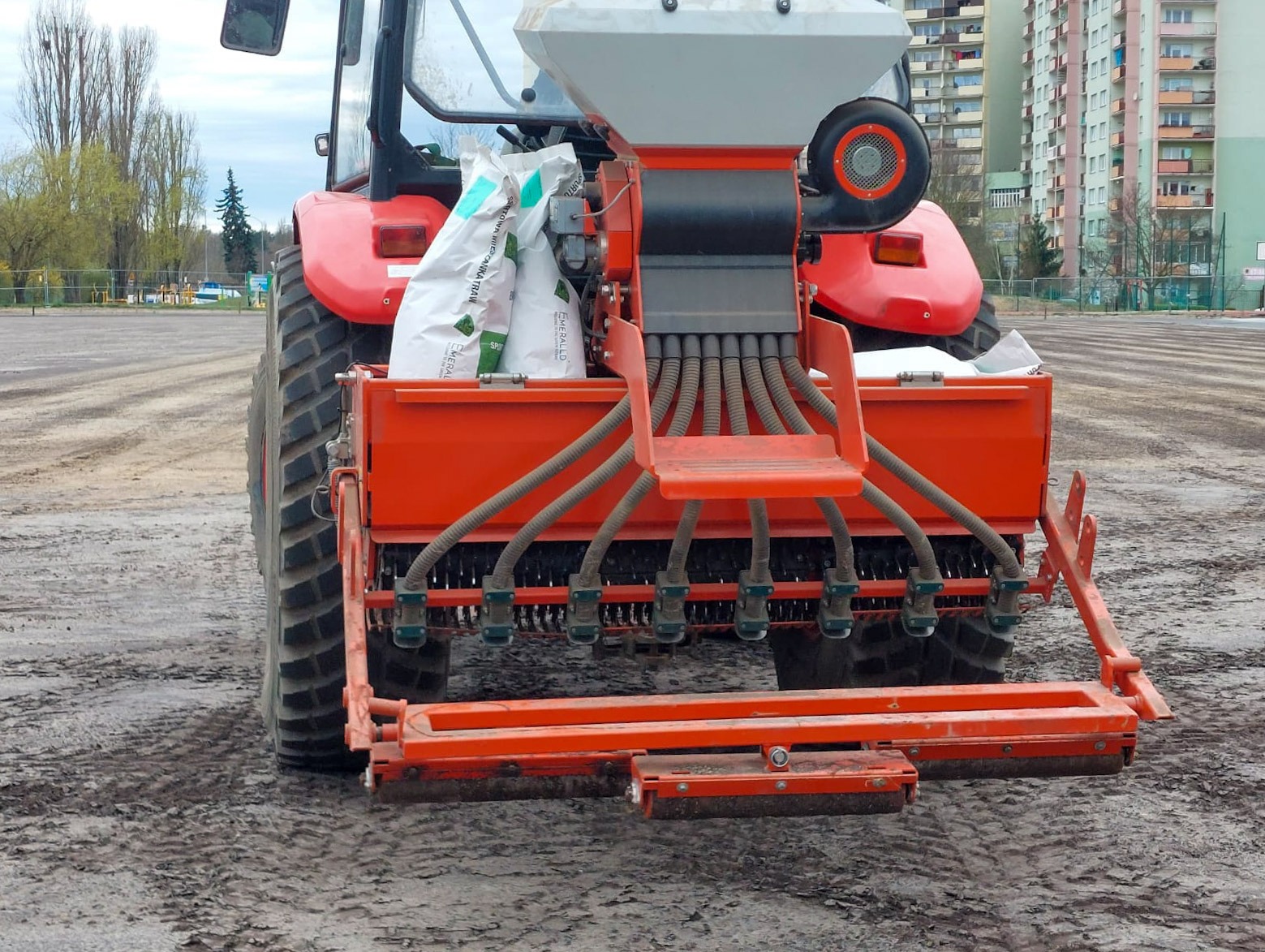 Czerwony traktor z siewnikiem punktowym na przygotowanym polu, w tle budynki mieszkalne.