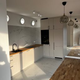 T-BUD - Nowoczesna kuchnia z białymi szafkami, blatem z jasnego drewna i marmurową ścianą, oświetlona wiszącymi lampami z przydymionego szkła, z widocznym przejściem do kolejnego pomieszczenia z parkietem.