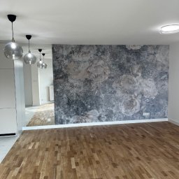 T-BUD - Pusty pokój z drewnianą podłogą parkietową, ścianą z fototapetą w kwiaty w odcieniach szarości i białymi ścianami, widoczne szklane lampy wiszące i odbicie pomieszczenia w lustrze.