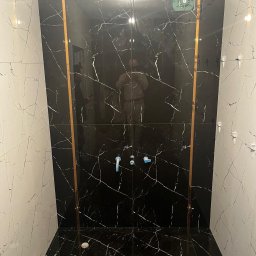 T-BUD - Wykończona łazienka z czarno-białymi płytkami imitującymi marmur, oddzielonymi złotymi listwami. Widoczne niebieskie zaślepki na rurach i wentylator sufitowy.