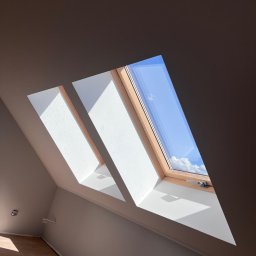 T-BUD - Poddasze z trzema oknami dachowymi, przez które wpada światło słoneczne. Widoczne fragmenty drewnianej podłogi i świeżo pomalowane ściany w odcieniach bieli i szarości.