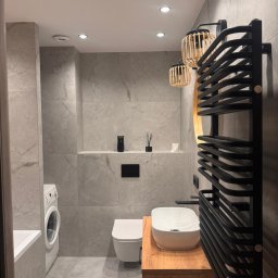 T-BUD - Nowoczesna łazienka z szarymi płytkami imitującymi marmur, drewnianą szafką pod umywalką i czarnym grzejnikiem drabinkowym. Widoczna pralka i toaleta.
