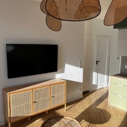 T-BUD - Jasne wnętrze po remoncie: parkiet w jodełkę, rattanowa komoda pod telewizorem, okrągły stolik kawowy i designerska lampa. Słoneczne refleksy na podłodze.