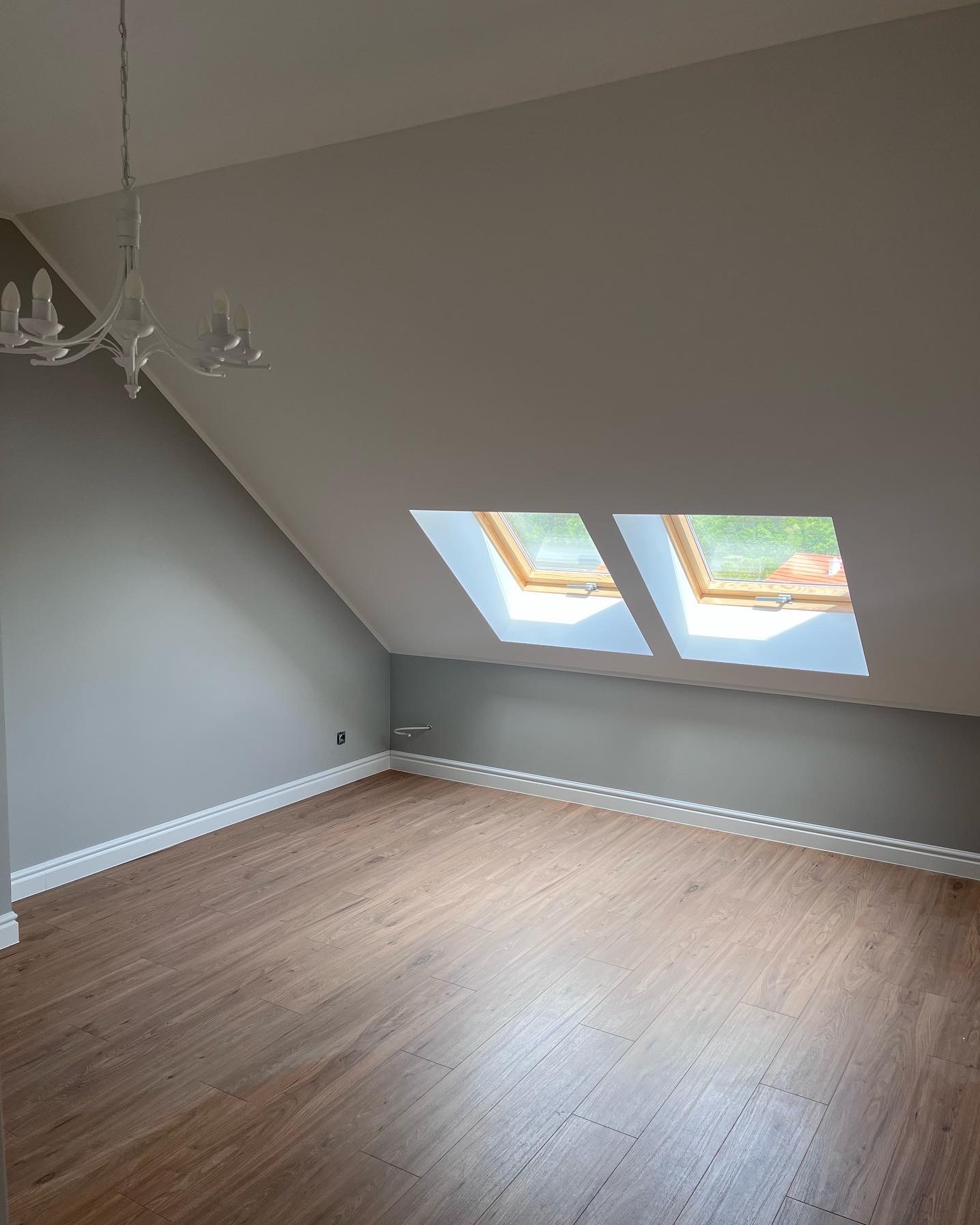 Poddasze po remoncie z drewnianą podłogą, szarą ścianą, białymi listwami przypodłogowymi oraz dwoma oknami dachowymi z widokiem na zieleń.