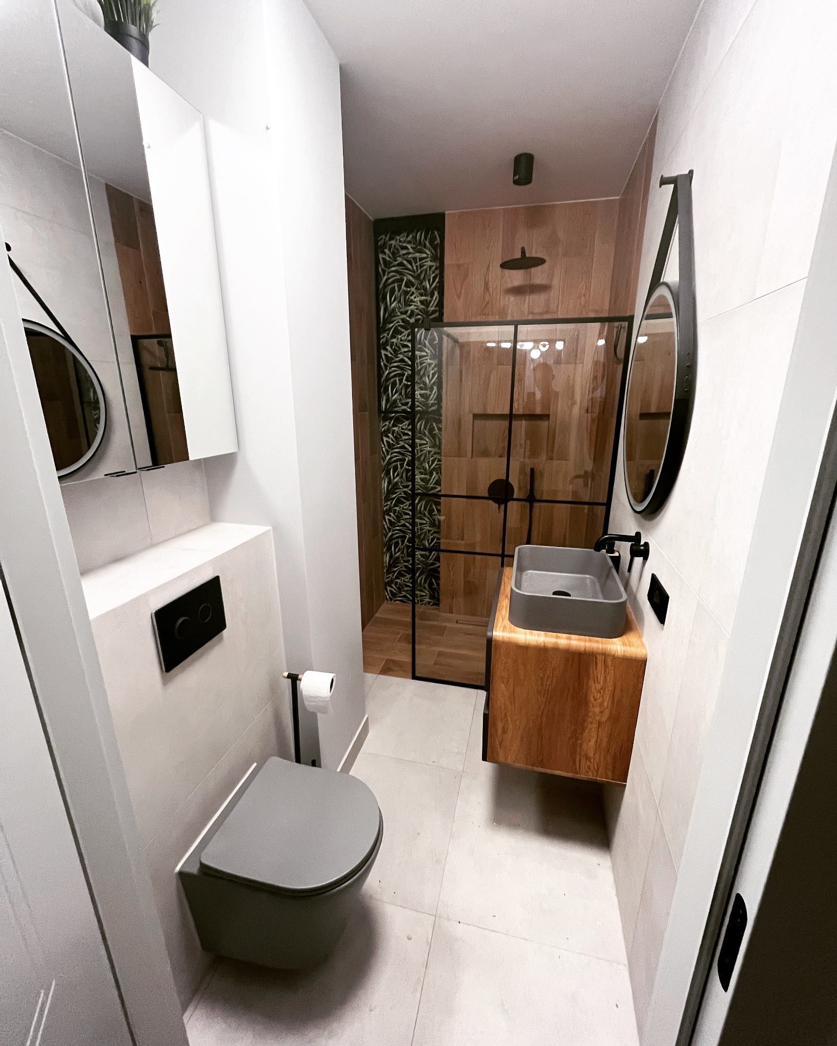 Nowoczesna łazienka z drewnianymi akcentami: kabina prysznicowa z czarną ramą, szara umywalka nablatowa na drewnianej szafce, wisząca toaleta w odcieniach szarości, okrągłe lustro z czarną ramą...