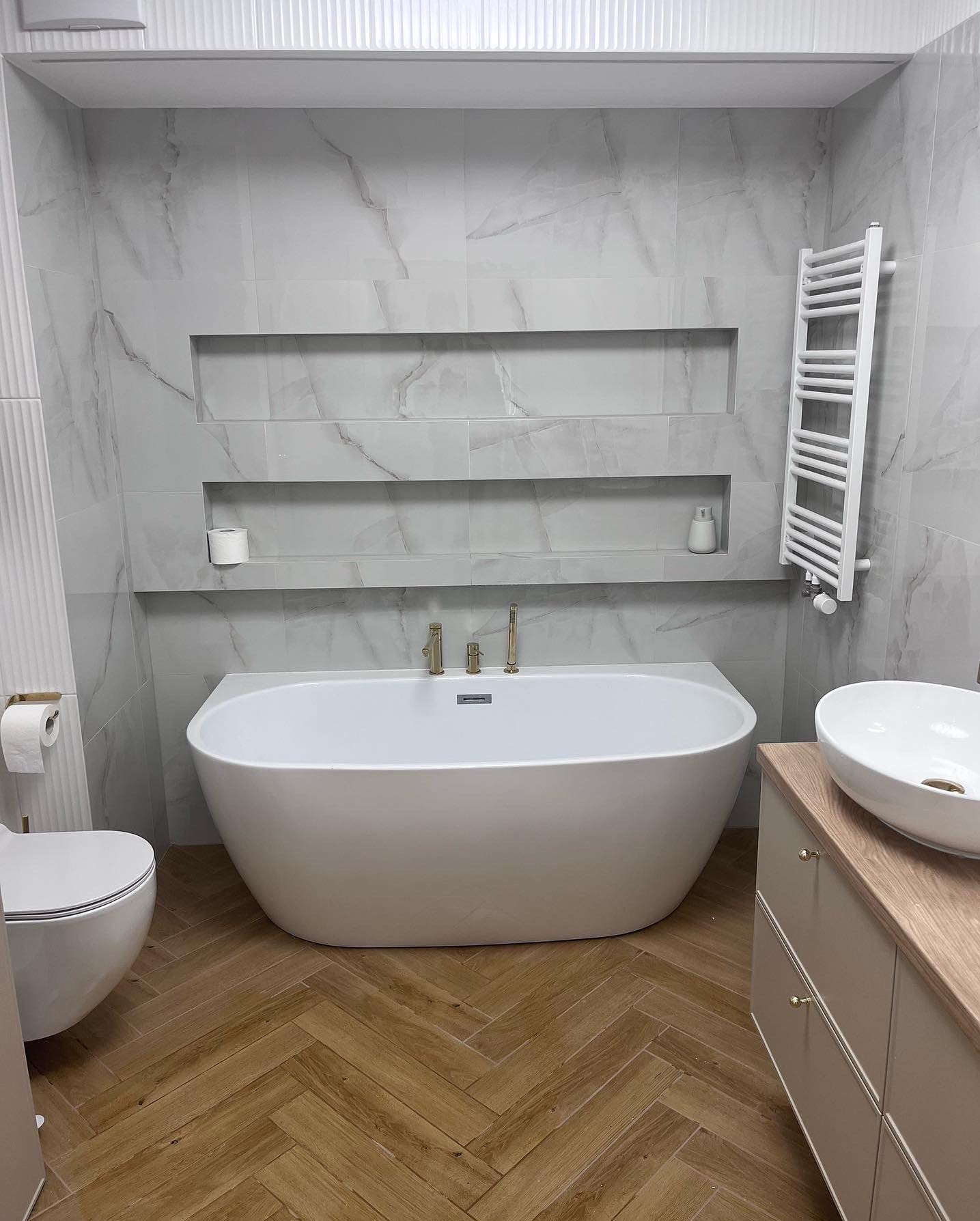 Nowoczesna łazienka z wolnostojącą wanną, marmurowymi płytkami ściennymi z wnękami, drewnianą podłogą ułożoną w jodełkę i białym grzejnikiem drabinkowym.
