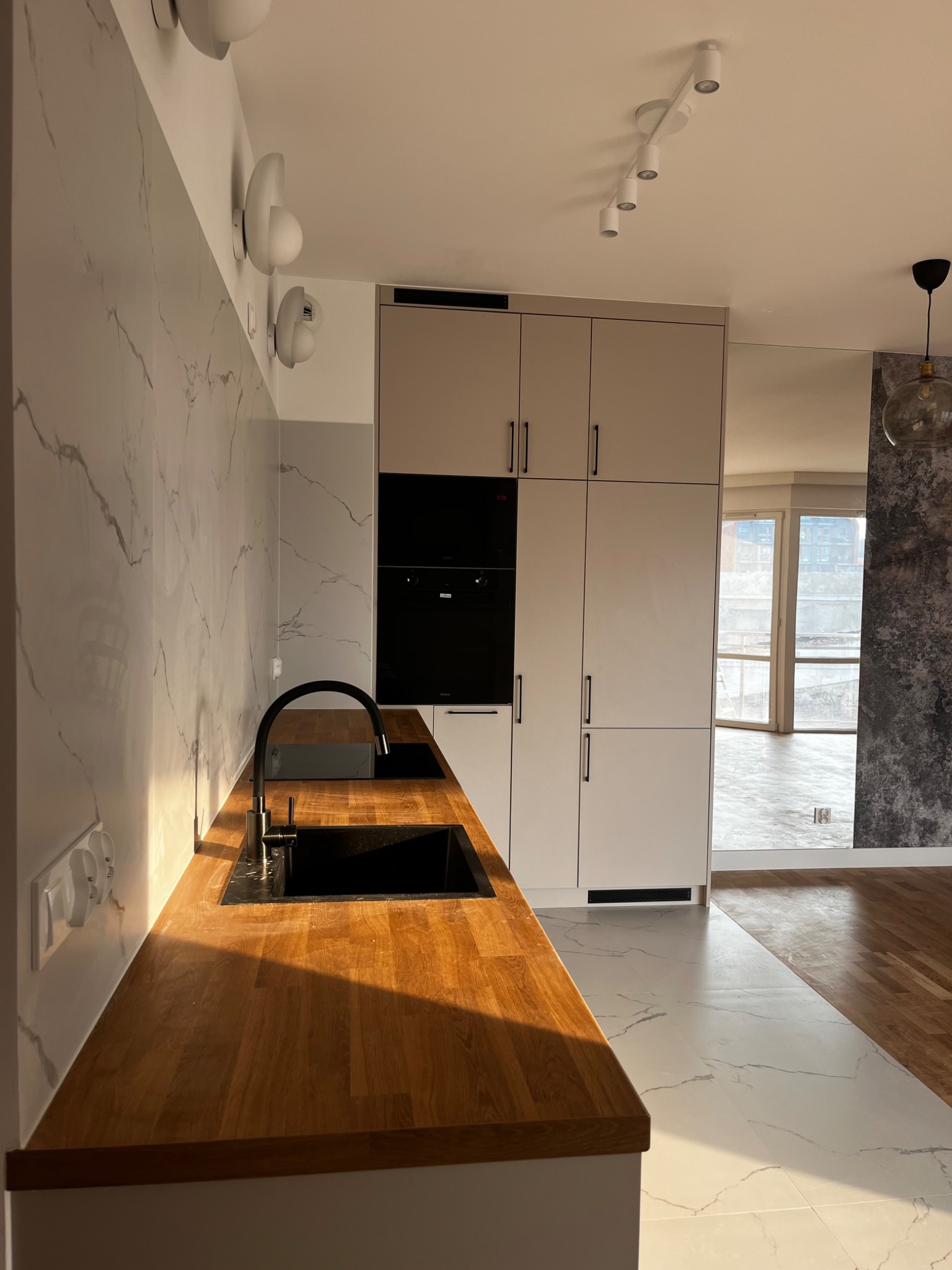 Nowoczesna kuchnia z drewnianym blatem, czarnym zlewem i baterią, marmurową ścianą oraz białymi szafkami, oświetlona ciepłym światłem słonecznym.