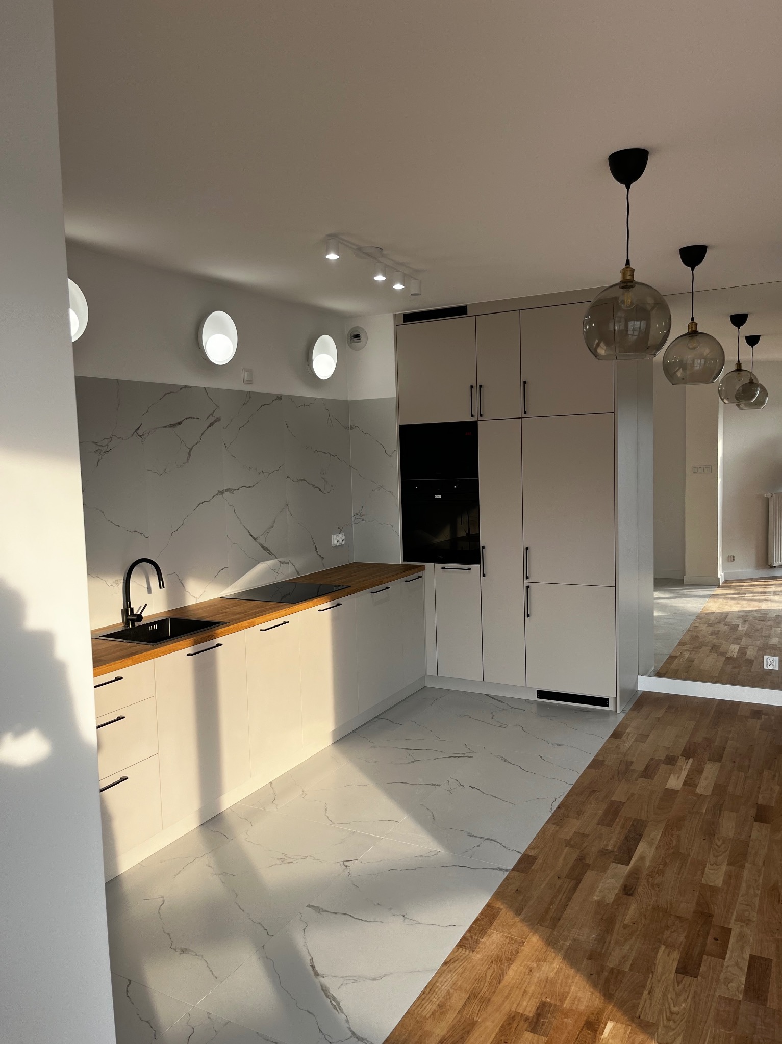 Nowoczesna kuchnia z białymi szafkami, blatem z jasnego drewna i marmurową ścianą, oświetlona wiszącymi lampami z przydymionego szkła, z widocznym przejściem do kolejnego pomieszczenia z parkietem.