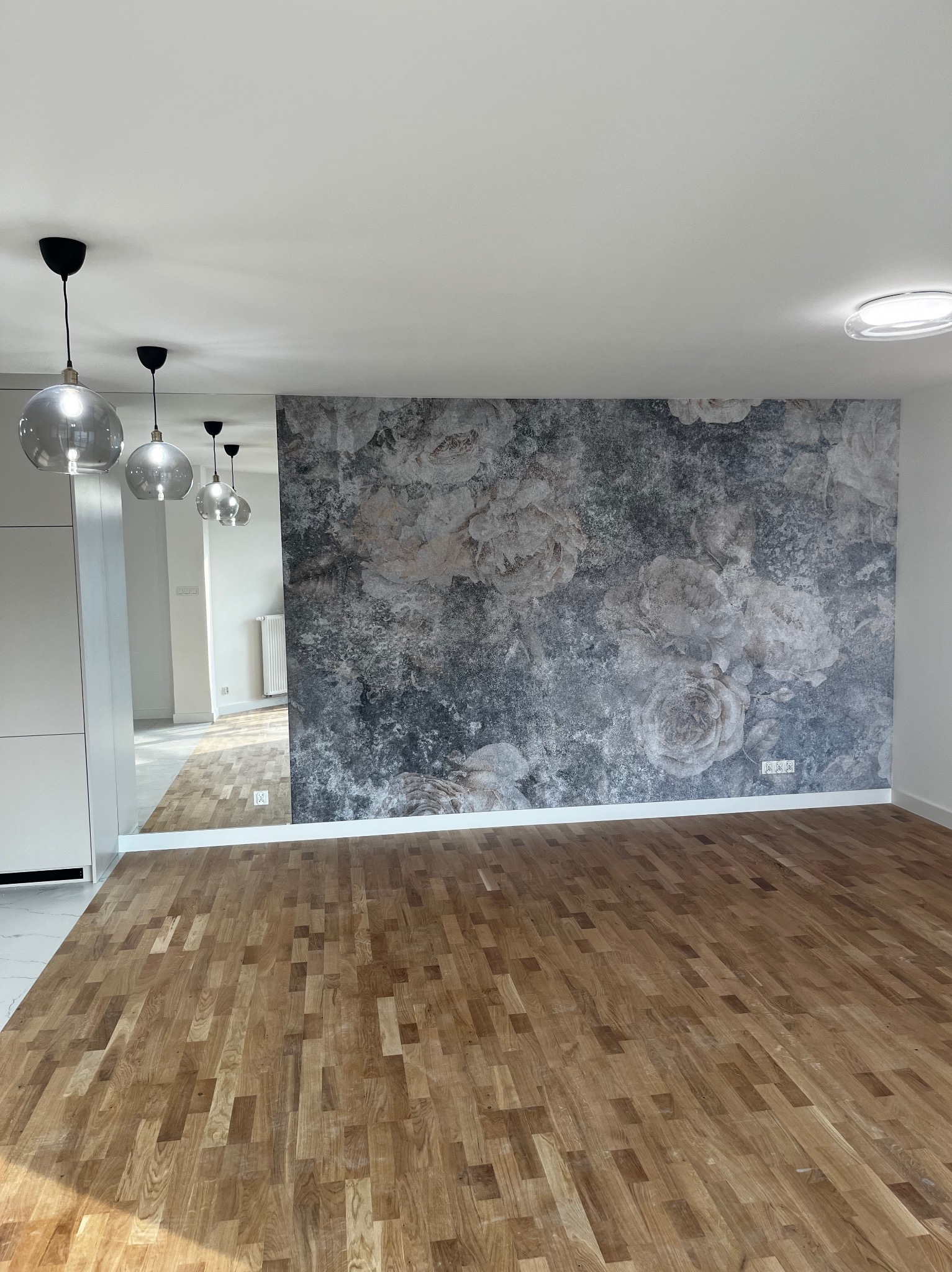 Pusty pokój z drewnianą podłogą parkietową, ścianą z fototapetą w kwiaty w odcieniach szarości i białymi ścianami, widoczne szklane lampy wiszące i odbicie pomieszczenia w lustrze.