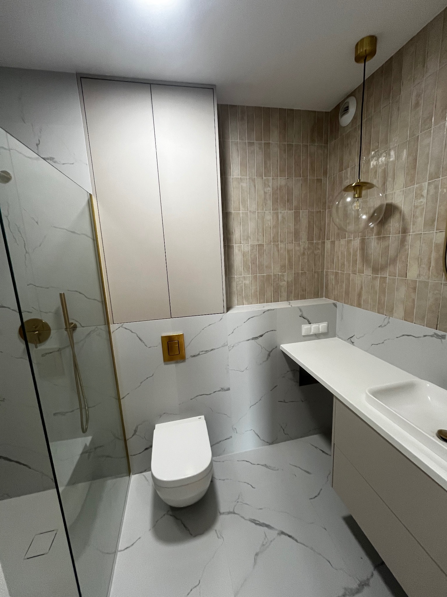 Nowoczesna łazienka z kabiną prysznicową ze złotymi detalami, podwieszaną toaletą, umywalką z szafką w odcieniach beżu oraz wiszącą lampą z przezroczystego szkła.