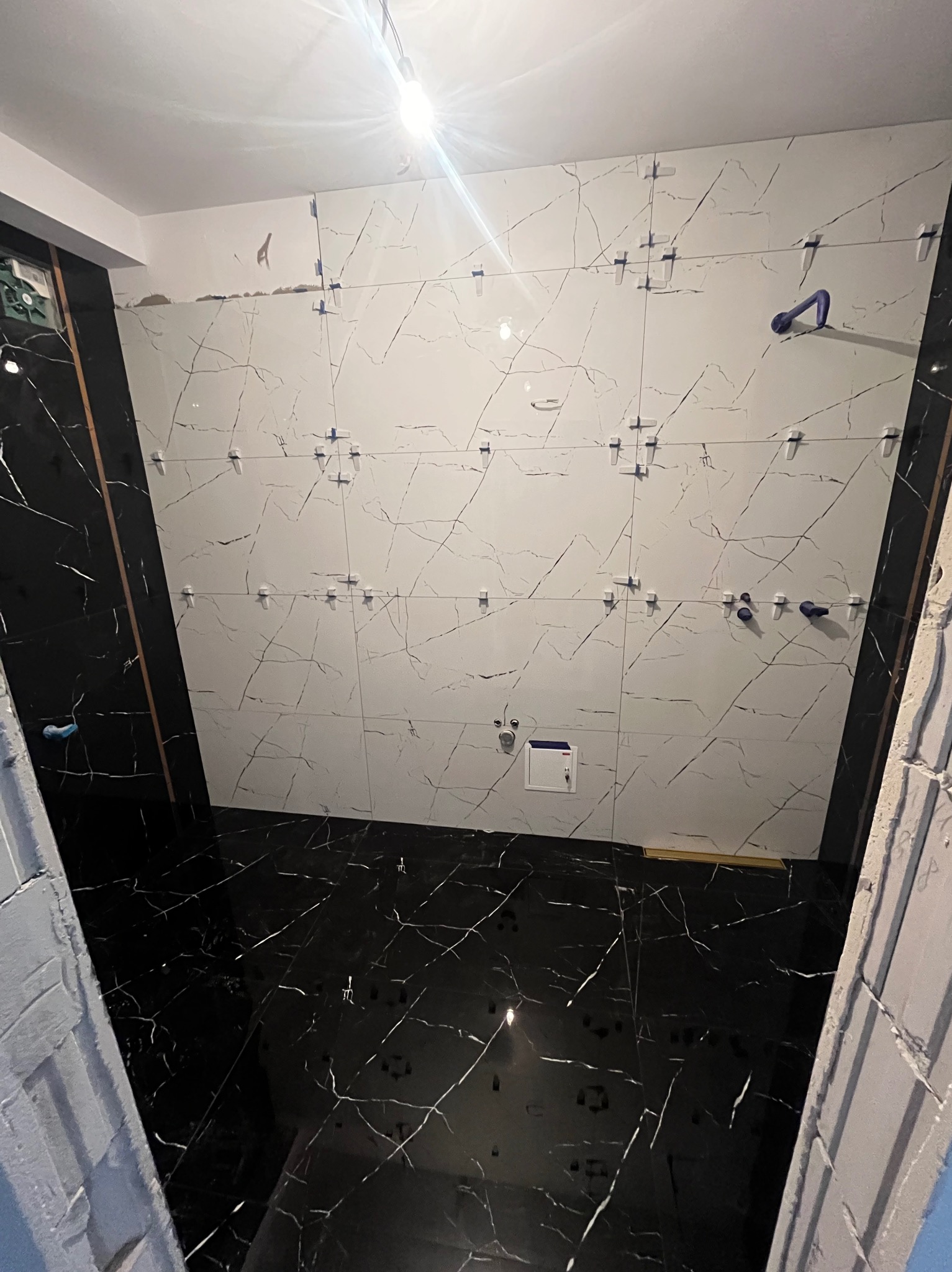 Łazienka w trakcie wykańczania, widoczne białe płytki imitujące marmur na ścianie z niebieskimi krzyżykami dystansowymi i czarne płytki na podłodze, również imitujące marmur.