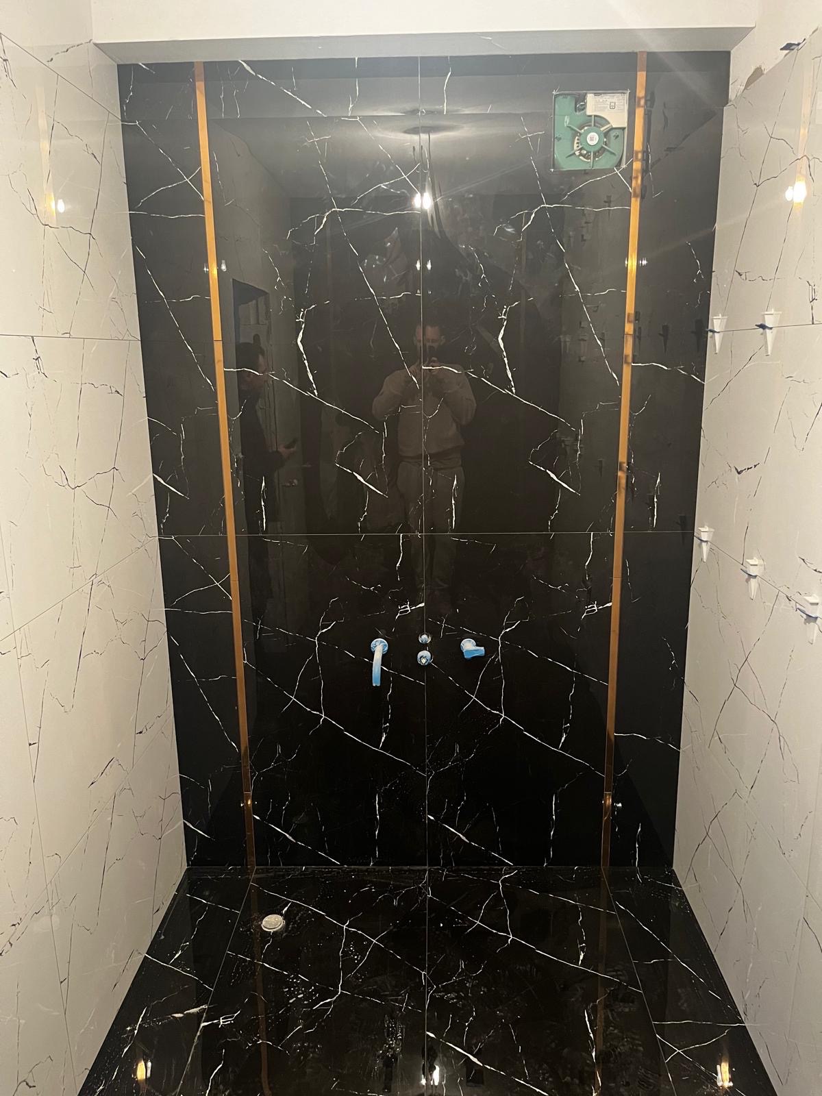 Wykończona łazienka z czarno-białymi płytkami imitującymi marmur, oddzielonymi złotymi listwami. Widoczne niebieskie zaślepki na rurach i wentylator sufitowy.