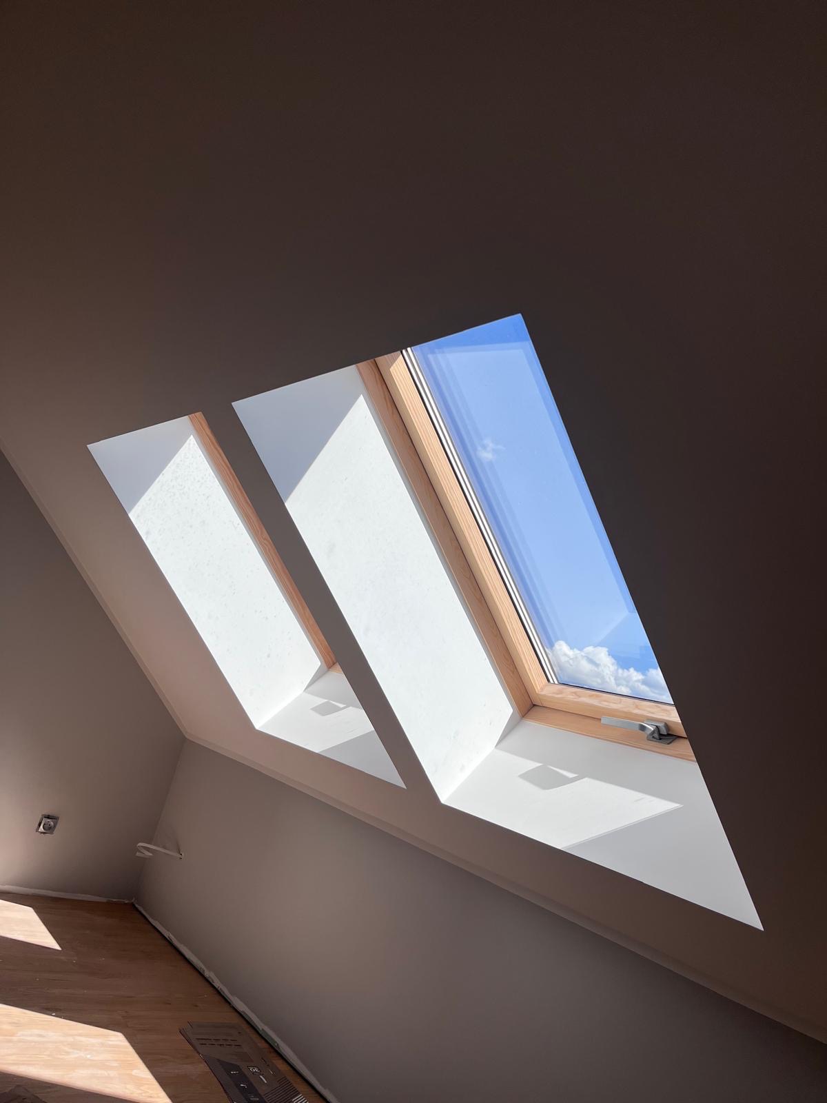 Poddasze z trzema oknami dachowymi, przez które wpada światło słoneczne. Widoczne fragmenty drewnianej podłogi i świeżo pomalowane ściany w odcieniach bieli i szarości.