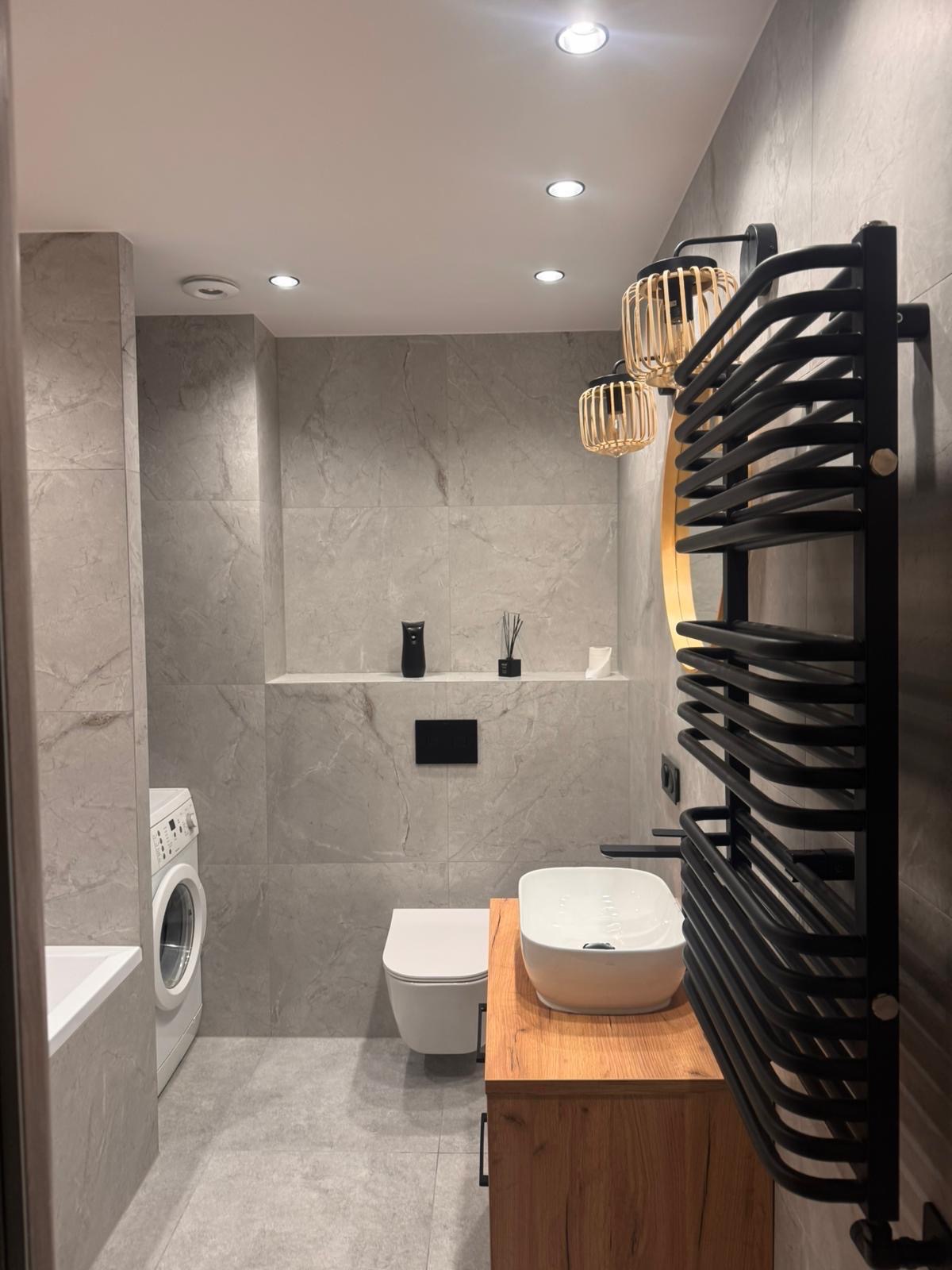 Nowoczesna łazienka z szarymi płytkami imitującymi marmur, drewnianą szafką pod umywalką i czarnym grzejnikiem drabinkowym. Widoczna pralka i toaleta.