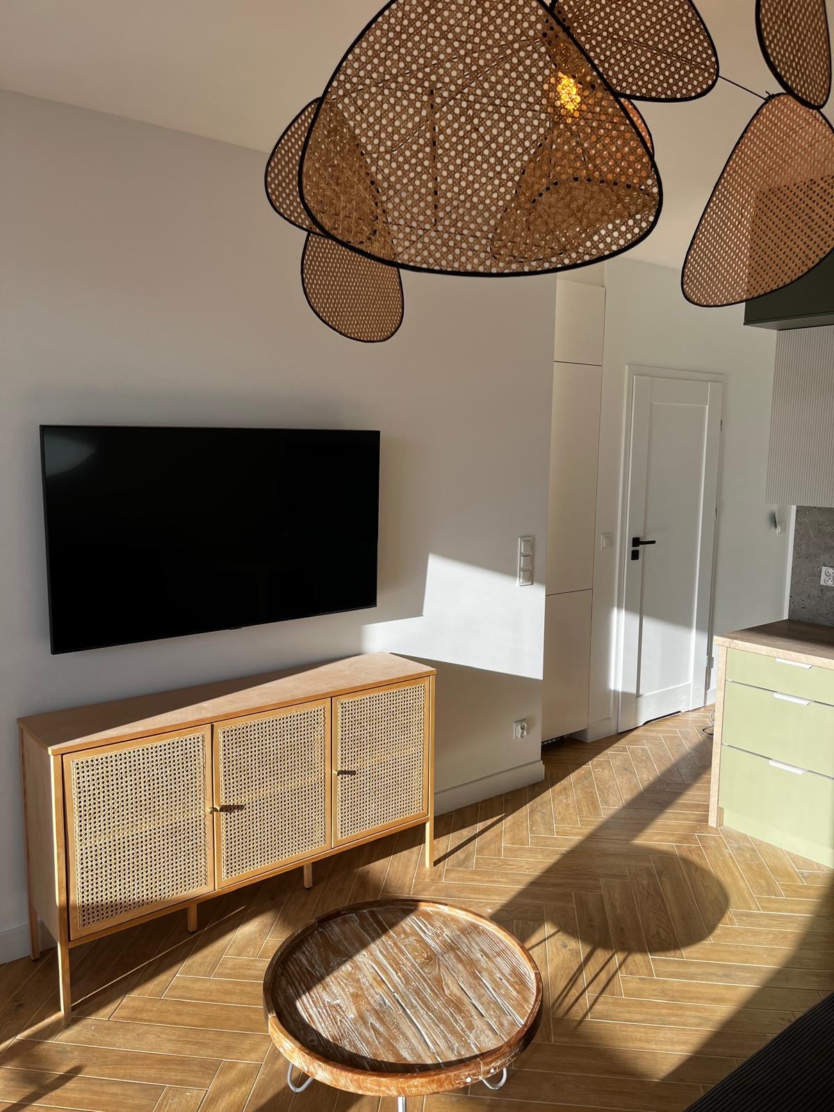 Jasne wnętrze po remoncie: parkiet w jodełkę, rattanowa komoda pod telewizorem, okrągły stolik kawowy i designerska lampa. Słoneczne refleksy na podłodze.