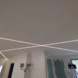 Wnętrze w trakcie remontu z zamontowanym oświetleniem LED w suficie, tworzącym geometryczny wzór krzyżujących się linii świetlnych, widoczne lampy budowlane na statywach.