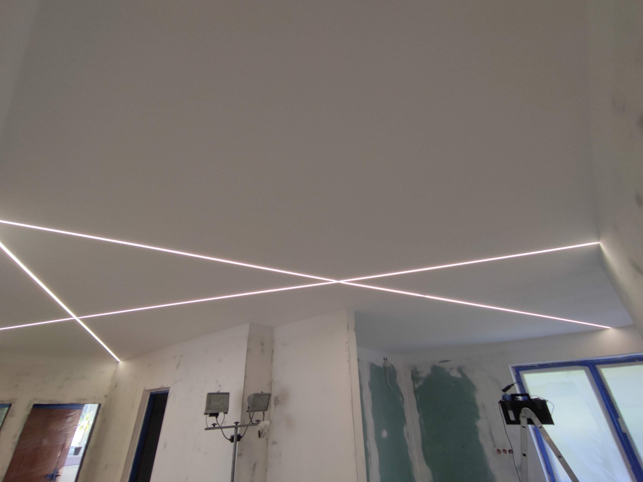 Wnętrze w trakcie remontu z zamontowanym oświetleniem LED w suficie, tworzącym geometryczny wzór krzyżujących się linii świetlnych, widoczne lampy budowlane na statywach.