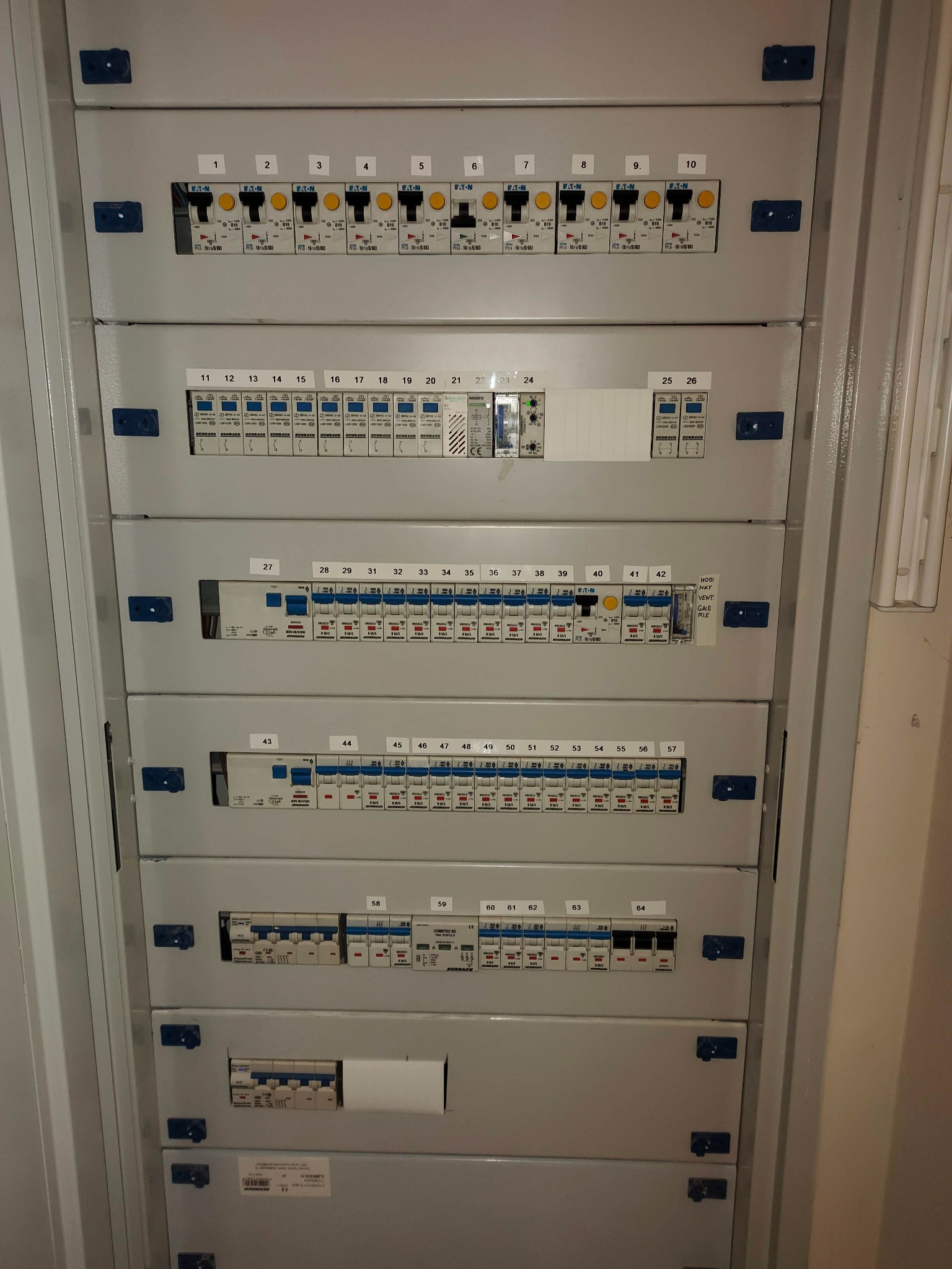 Rozdzielnica elektryczna z licznymi bezpiecznikami i wyłącznikami różnicowoprądowymi, oznaczona numerami od 1 do 64, widoczne różne marki i modele zabezpieczeń.