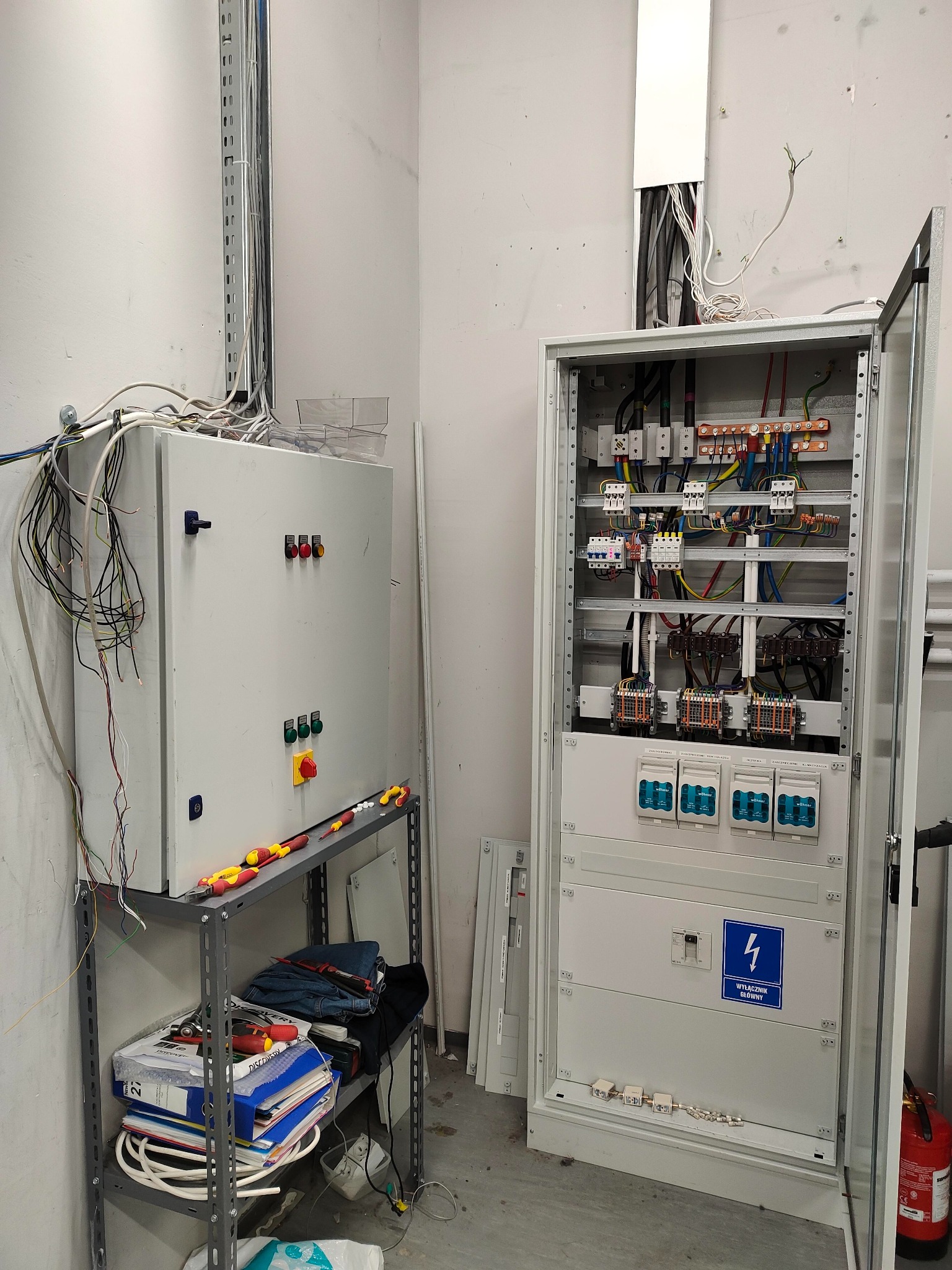Otwarta szafa elektryczna z widocznymi bezpiecznikami, przewodami i elementami sterującymi, obok druga szafa z przyciskami i przełącznikami, na metalowym regale leżą narzędzia i dokumenty.