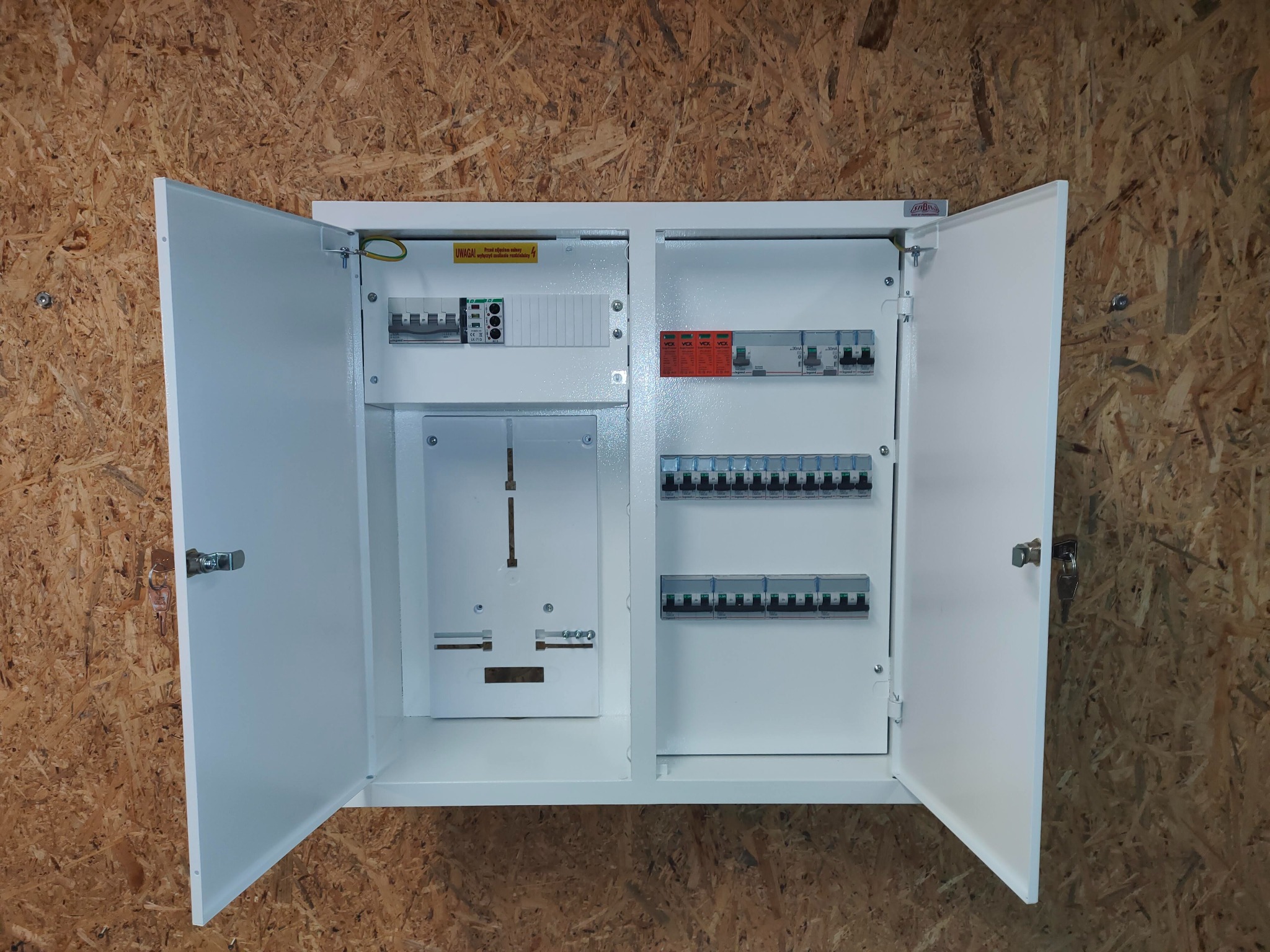 Otwarta, biała skrzynka elektryczna z zainstalowanymi bezpiecznikami i elementami montażowymi, zamontowana na ścianie z płyty OSB.