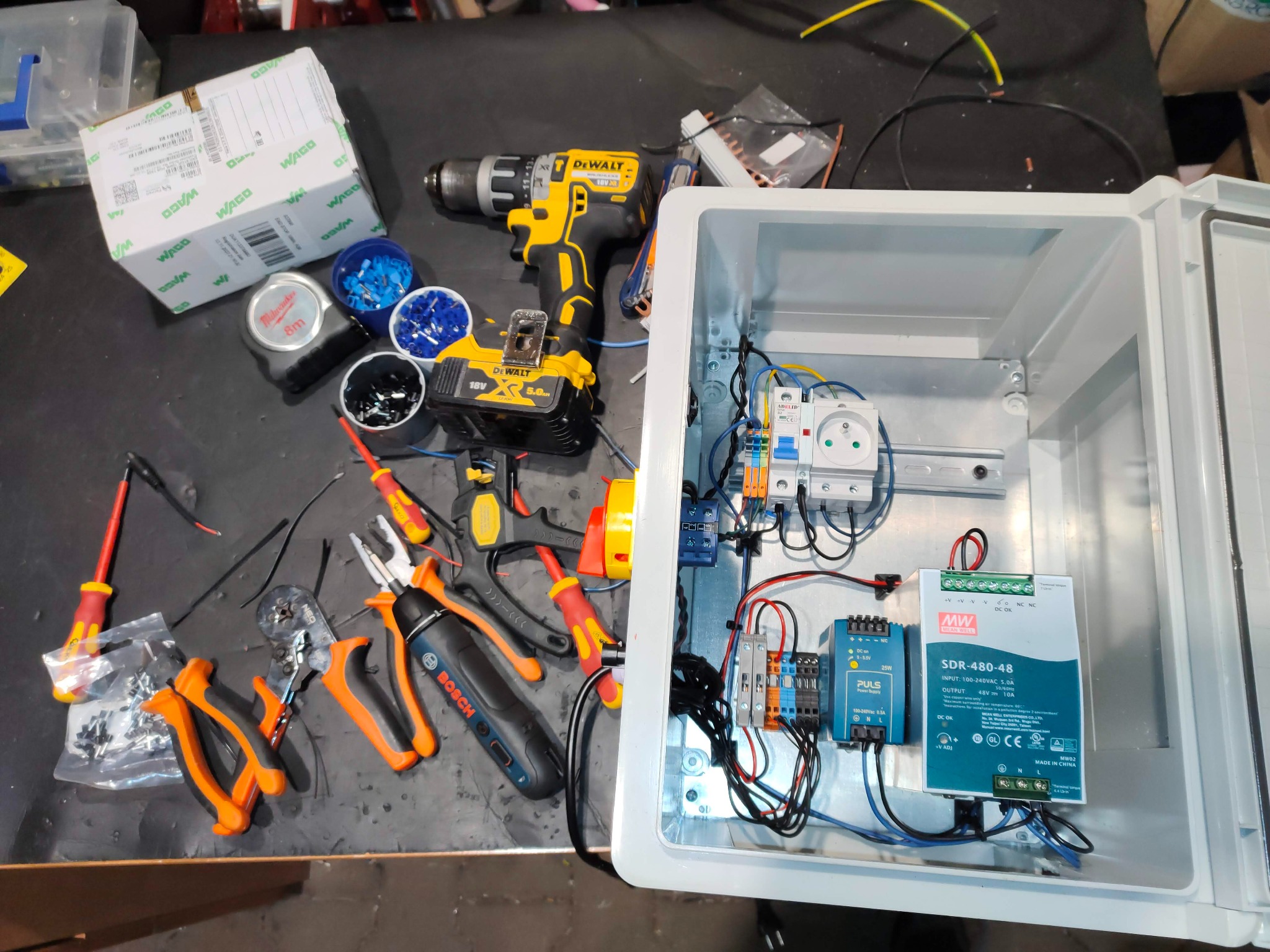 Panel elektryczny w trakcie montażu: widoczne narzędzia (wiertarka DeWalt, kombinerki Bosch, miernik Milwaukee), przewody, zasilacz Mean Well SDR-480-48 i Puls 25W, oraz komponenty WAGO. Czarna...