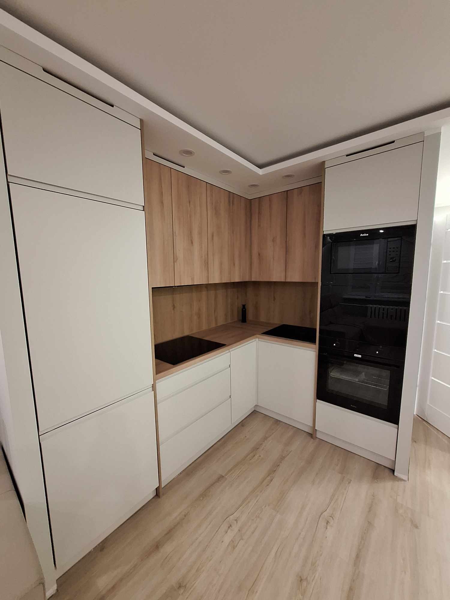 Nowoczesna kuchnia na wymiar: białe fronty szafek dolnych i górnych, drewnopodobne szafki wiszące, czarny piekarnik i płyta indukcyjna Amica, jasna podłoga.